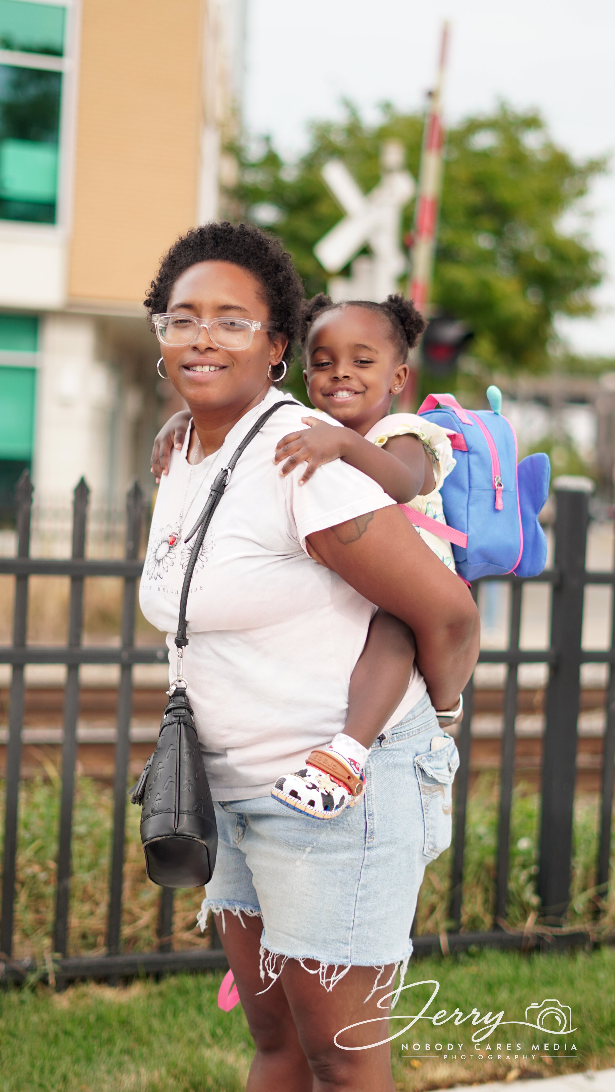 A woman carrying a young girl on her back outdoors with a fence and train crossing sign in the background.