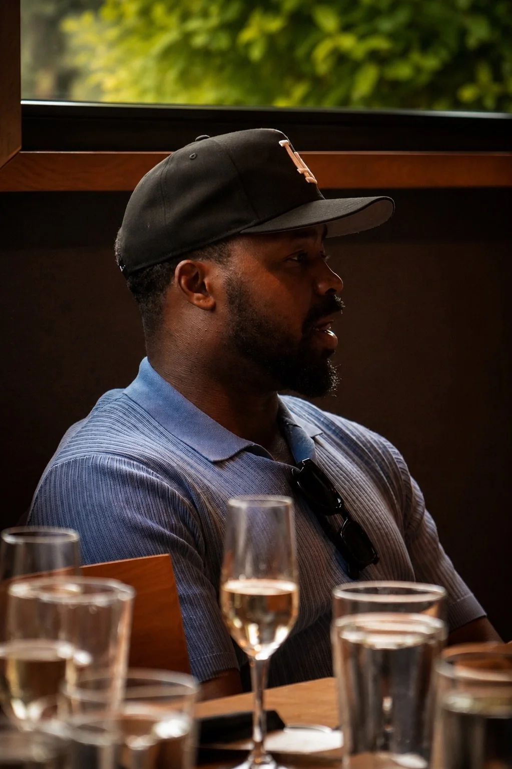 A man with dark skin, short hair, and a beard, wearing a black baseball cap and a light blue collared shirt, sitting at a table in a restaurant or bar, with glasses of champagne or sparkling wine in front of him. The background shows a window with green foliage outside.