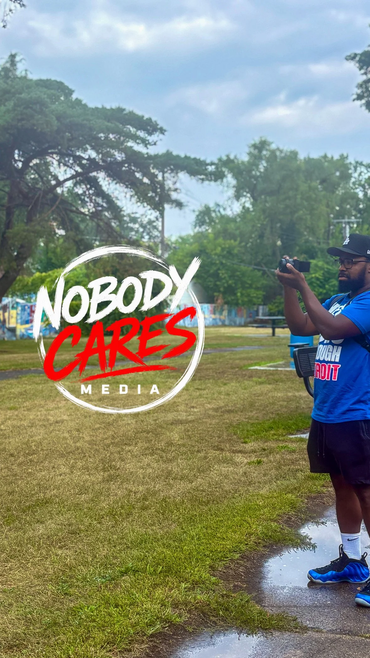 A man in a blue shirt, black shorts, and blue sneakers stands on a muddy patch of grass at a park, holding a phone camera. The background shows trees, cloudy sky, and a graffiti-covered wall. Overlaid text reads 'Nobody Cares Media'.