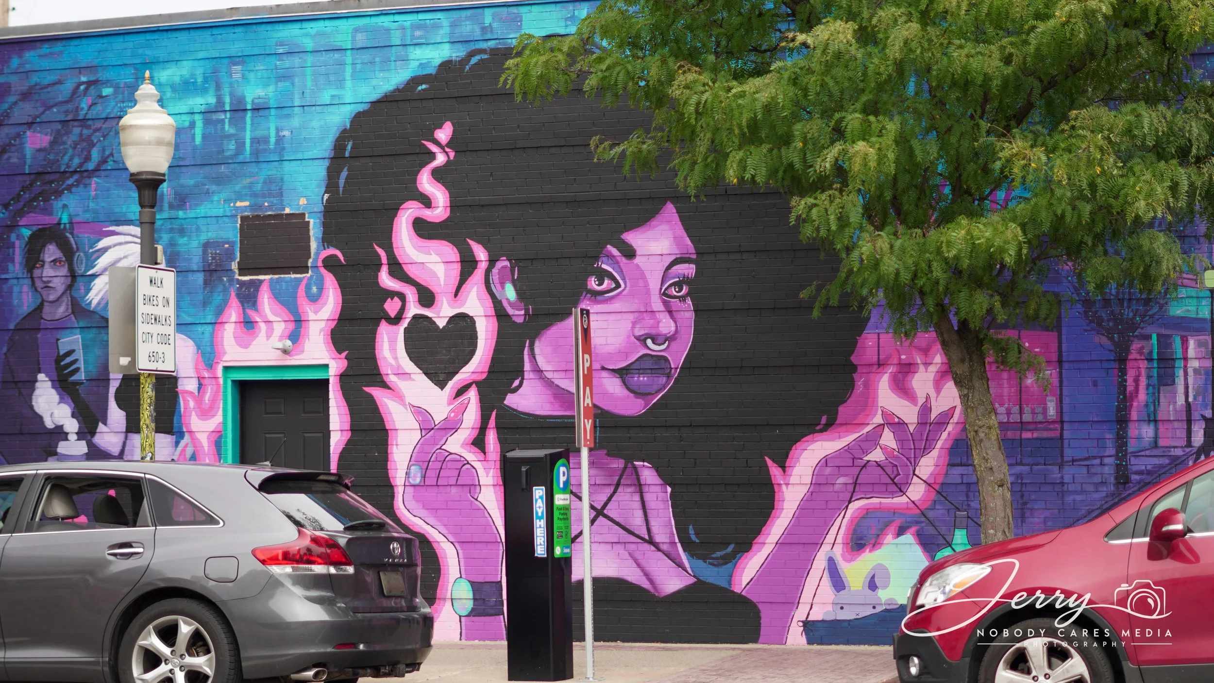 Colorful mural on a brick wall depicting a woman with dark hair, purple skin, and expressive eyes, surrounded by pink flames and abstract cityscape background, with cars parked in front.