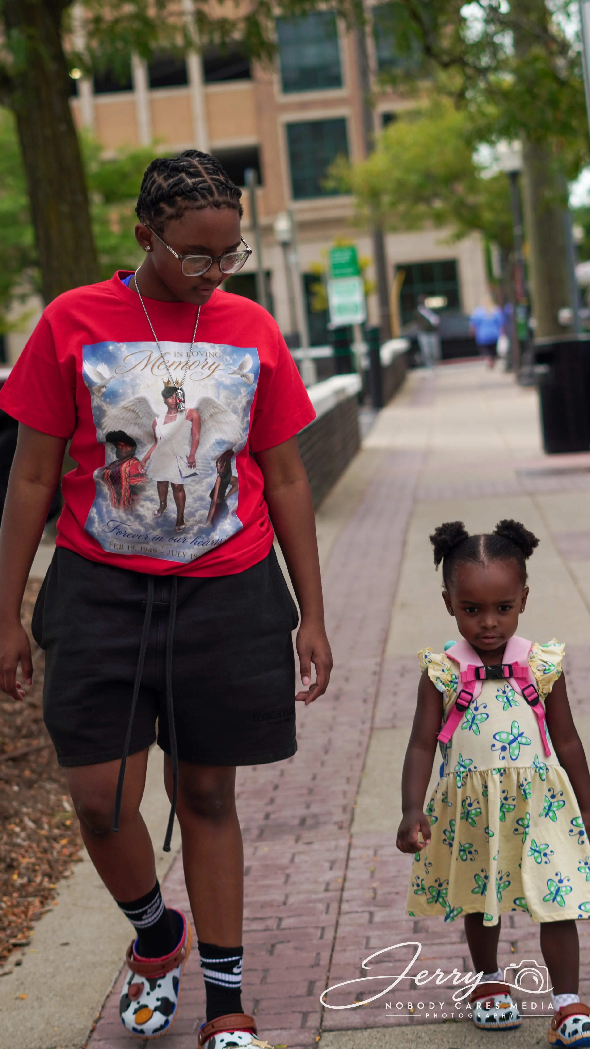 A woman and a young girl walking on a sidewalk in an urban area with trees and buildings in the background. The woman wears glasses, a red t-shirt with a memorial design, black shorts, and cow-themed sneakers. The girl wears a yellow dress with a butterfly pattern, a pink backpack, and sandals.