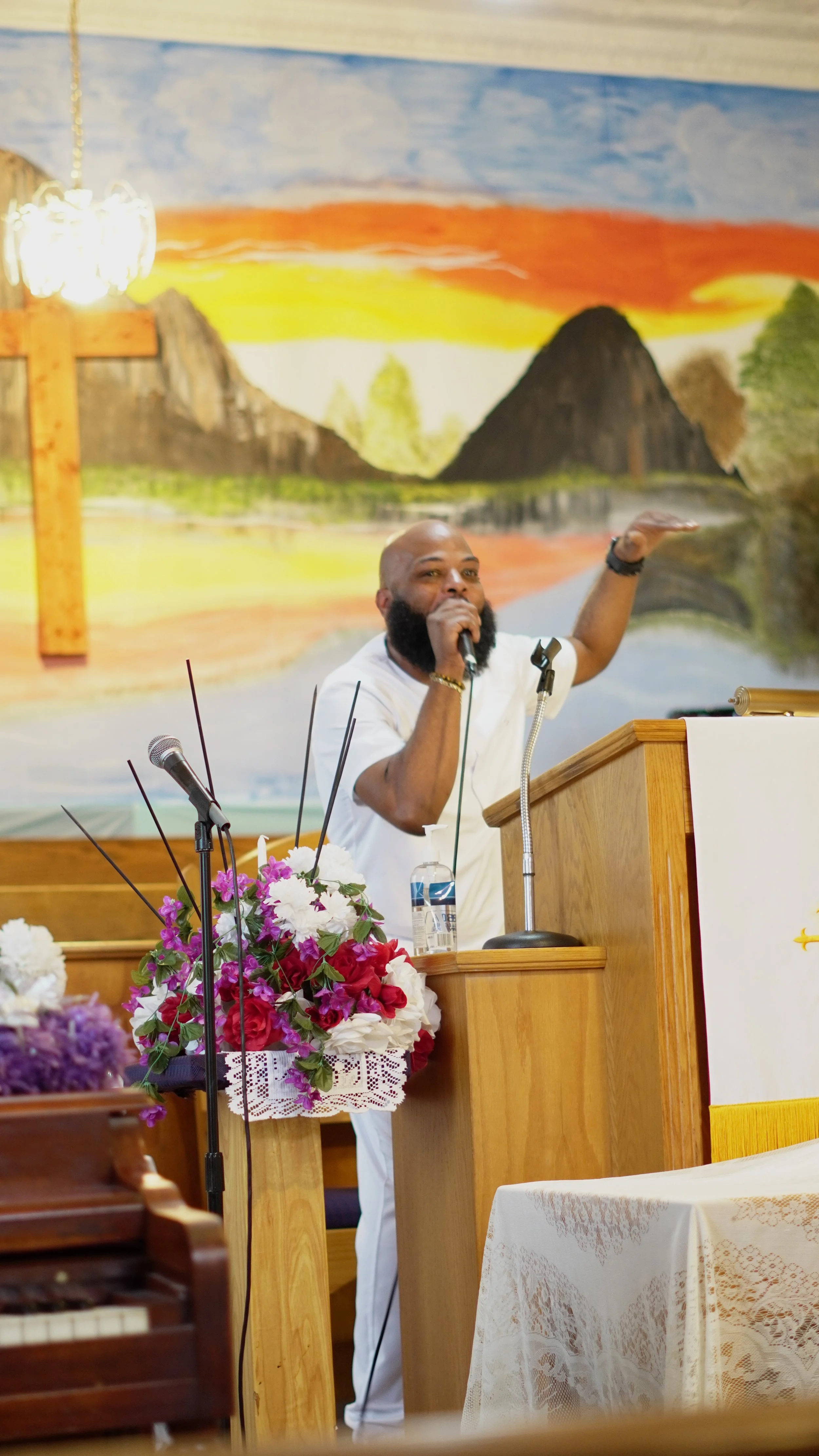 A man with a beard and white shirt speaking or singing into a microphone from a wooden pulpit in a church decorated with flowers, with a painted mountain and sunset scene in the background.