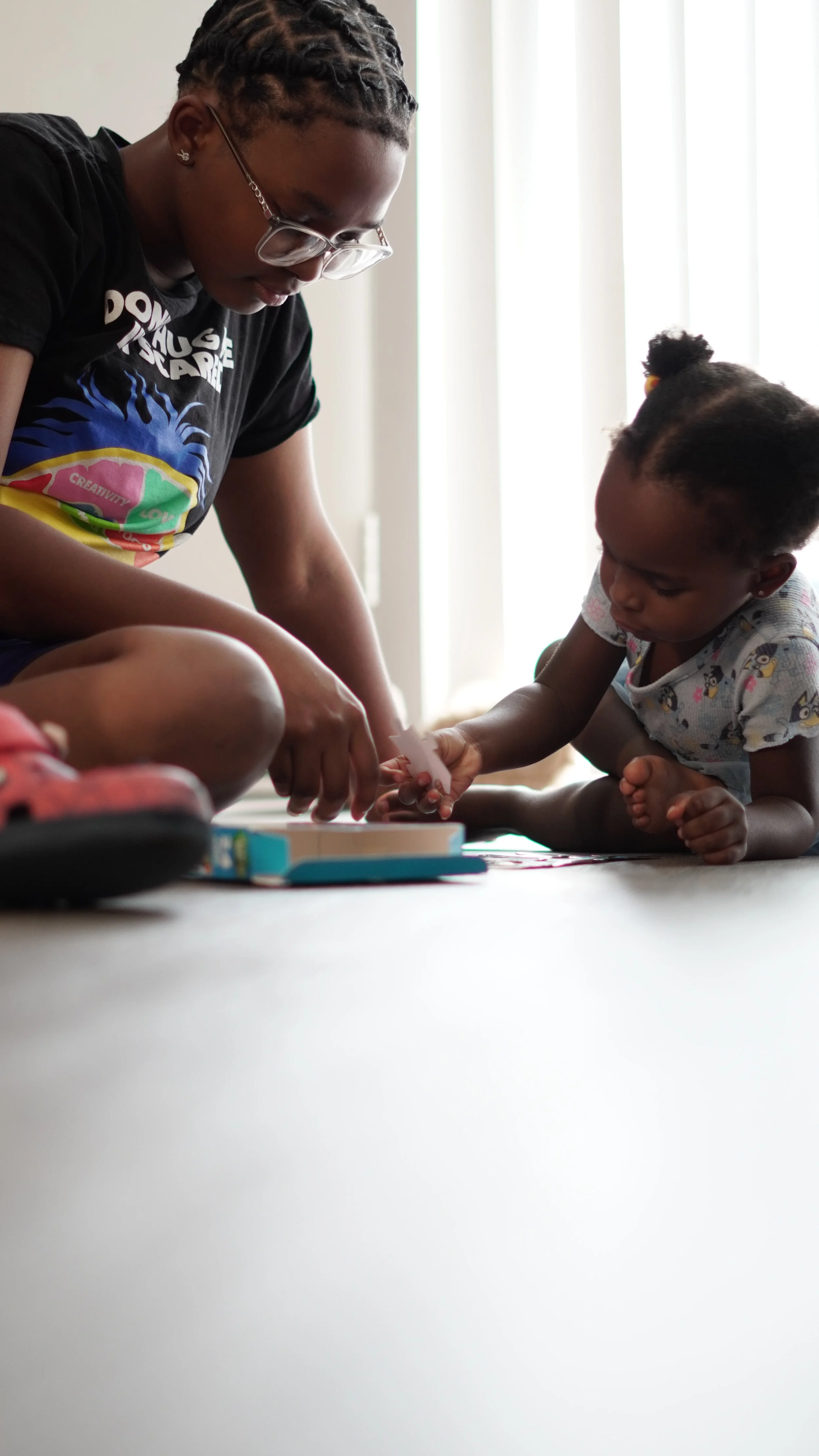 A young woman and a toddler girl are sitting on the floor, engaged in a shared activity involving small objects on a game box, near a bright window.