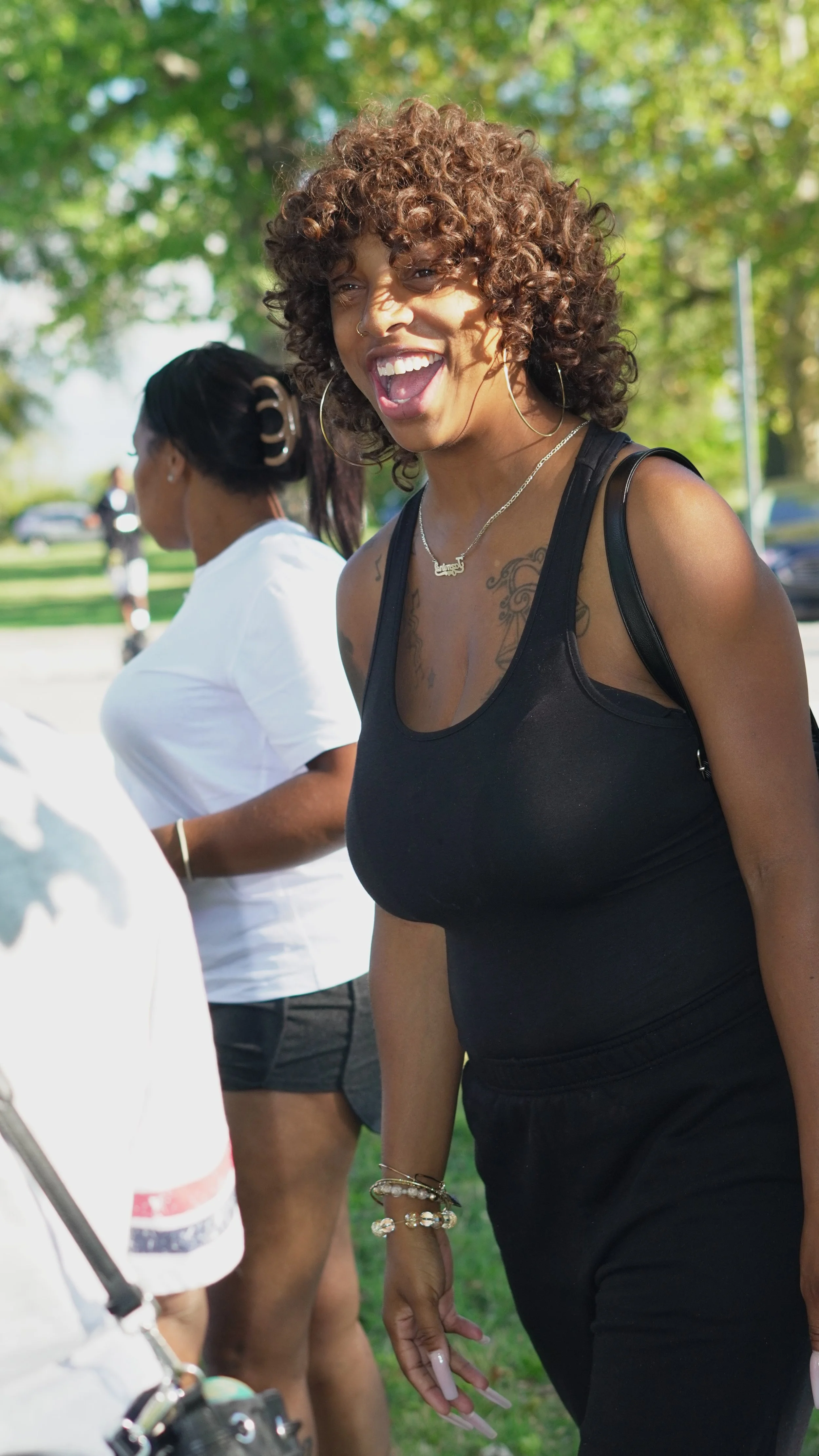 A woman with curly hair, wearing a black top and jewelry, smiling outdoors in a park with trees in the background.