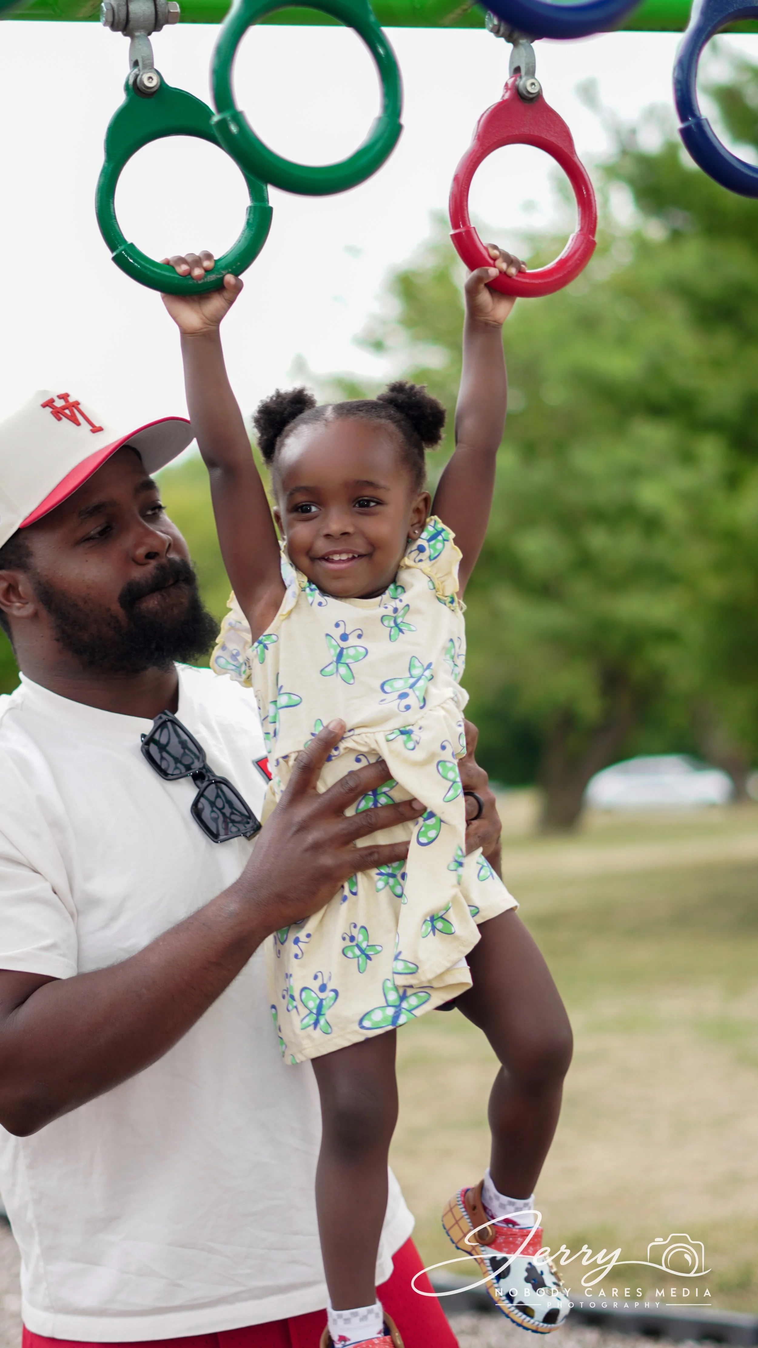 A man holding up a young girl by her waist at a park. The girl is hanging from playground rings, smiling, wearing a yellow dress with butterfly prints and colorful sneakers. The man is wearing a white shirt, red shorts, and a white baseball cap.