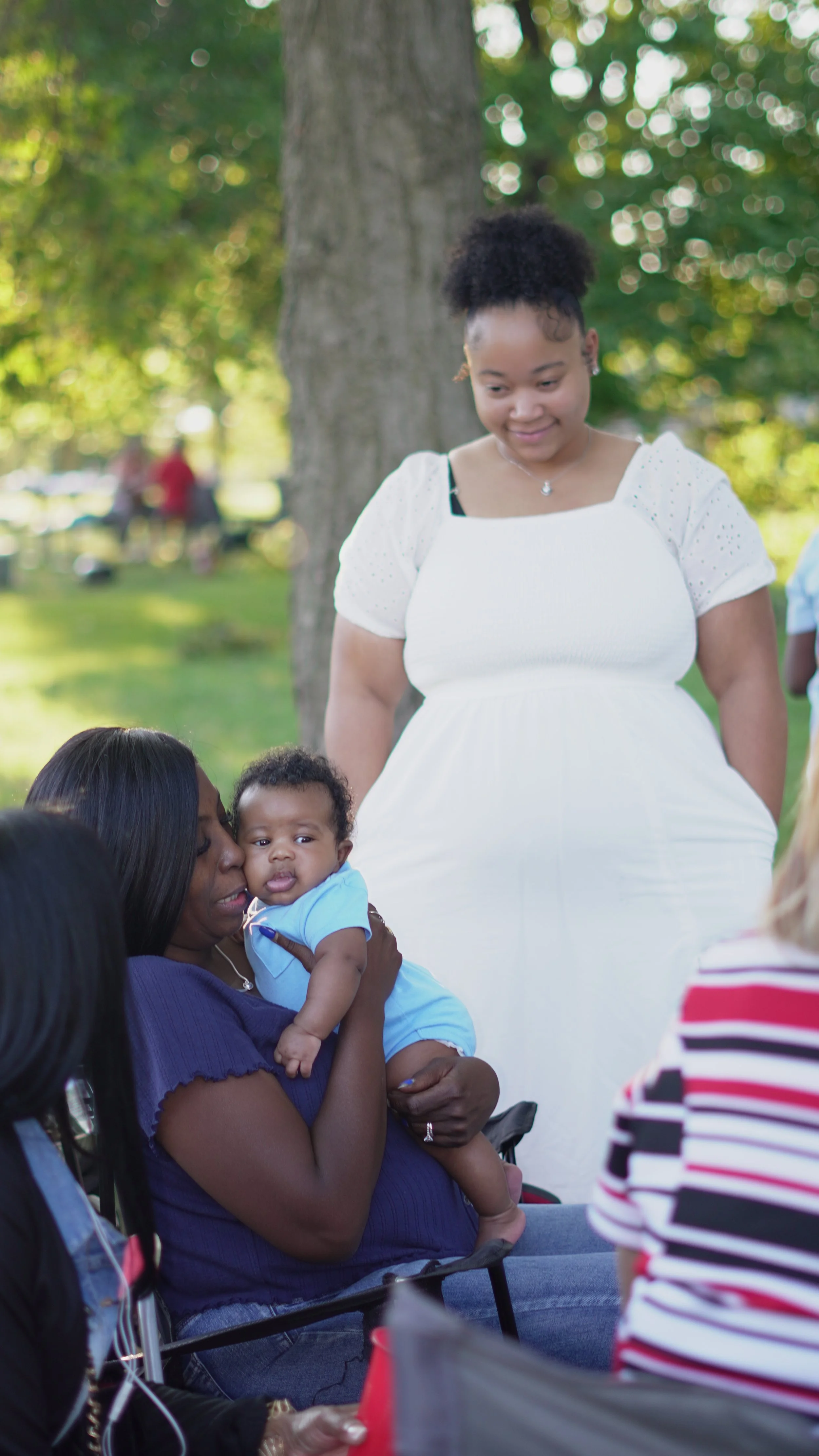 A woman with dark hair holding a baby, while a woman with short curly hair in a white dress stands behind them outdoors near a tree, in a park on a sunny day.