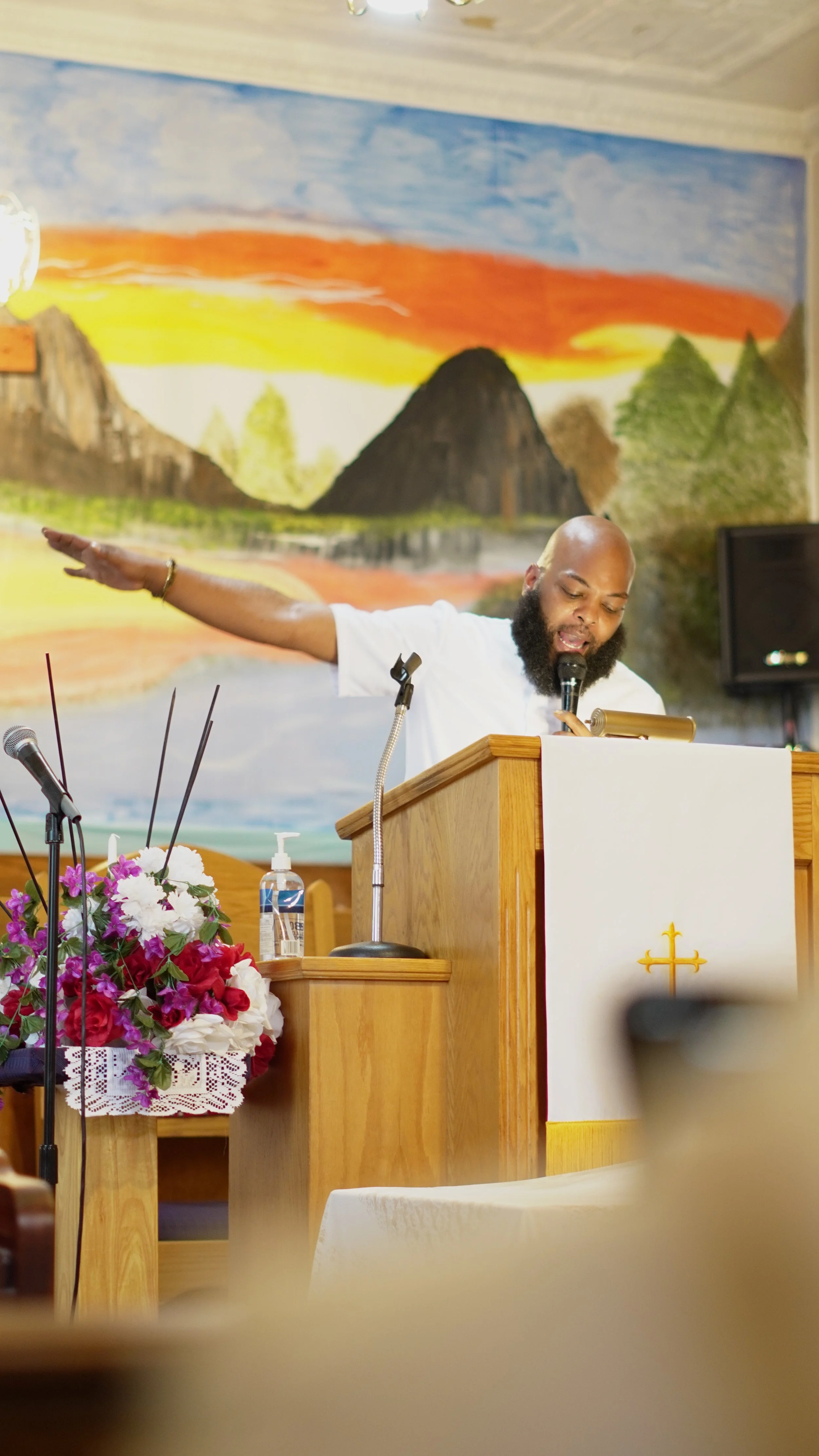 A man with a beard and bald head standing at a pulpit inside a church, wearing a white shirt, with his eyes closed and one arm extended outward. The pulpit has a white cloth with a gold cross, and there are flowers and hand sanitizer on a table nearby. A colorful mountain and sunset mural decorates the background wall.