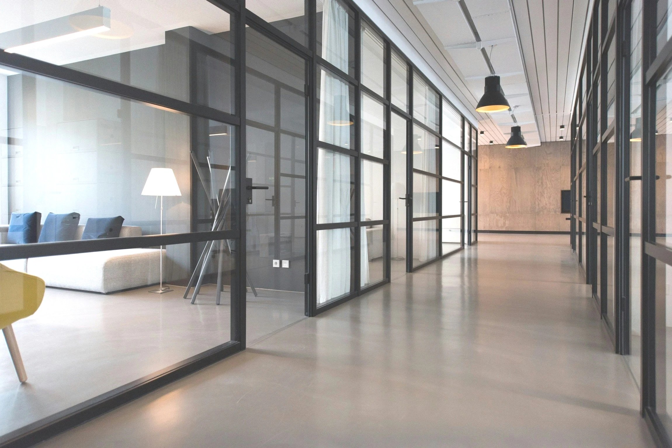 Innenaufnahme eines modernen Büros mit viel Glas, einem Sofa, einem Stehlampe und Pendelleuchten an der Decke.