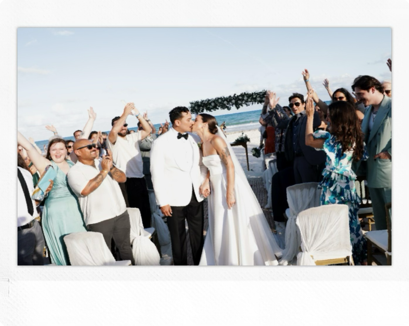 Pareja de novios besándose en una ceremonia de boda en la playa, rodeados de invitados.