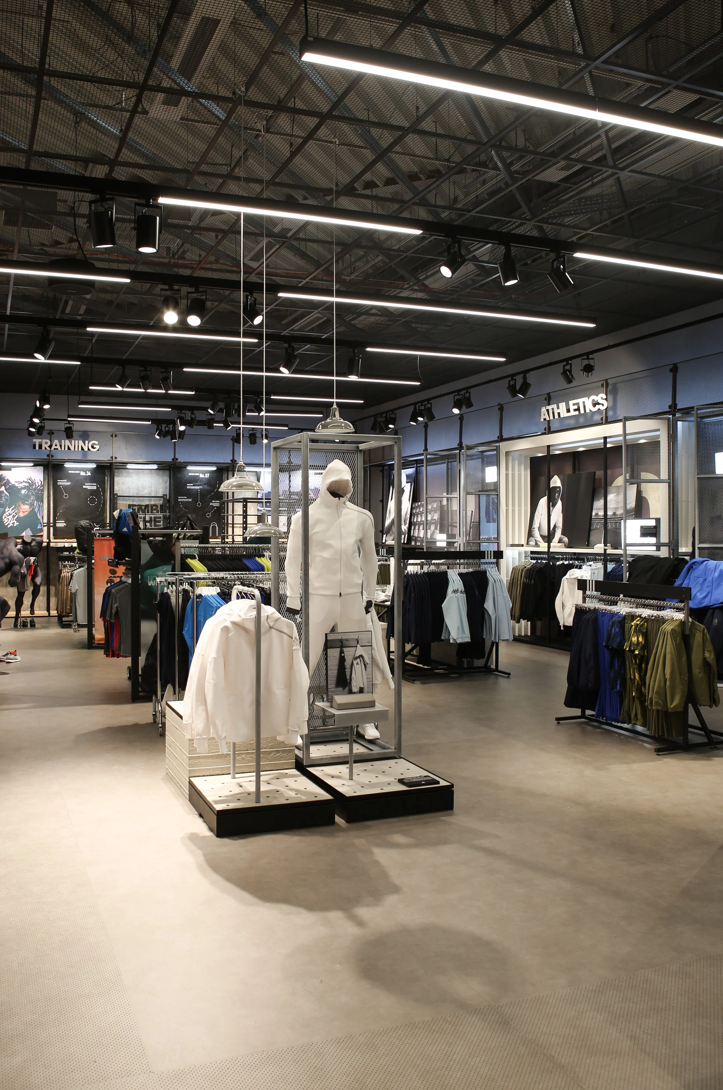Interior of a sportswear store with display racks of athletic clothing and mannequins, shelves labeled 'Training' and 'Athletics', and industrial ceiling with exposed grid and lighting fixtures.
