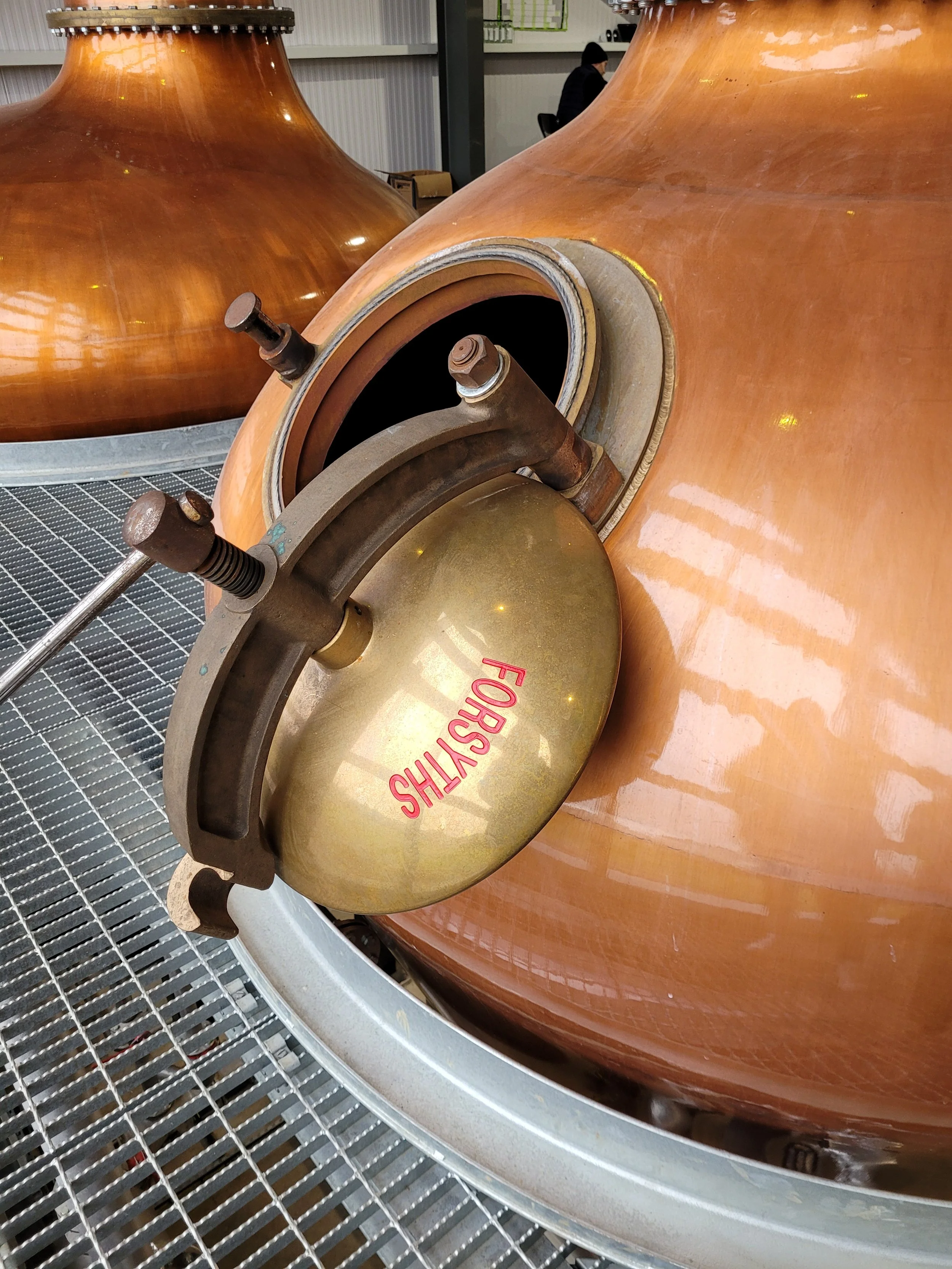 Close-up of a copper distillation still with a view of the opening and fitting, labeled 'FORSYTH' on the side, situated on a metal grate in an industrial or manufacturing setting.