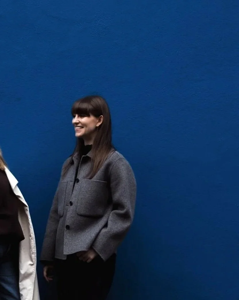 Woman with long dark hair and bangs smiling, wearing a grey jacket, standing against a blue wall.