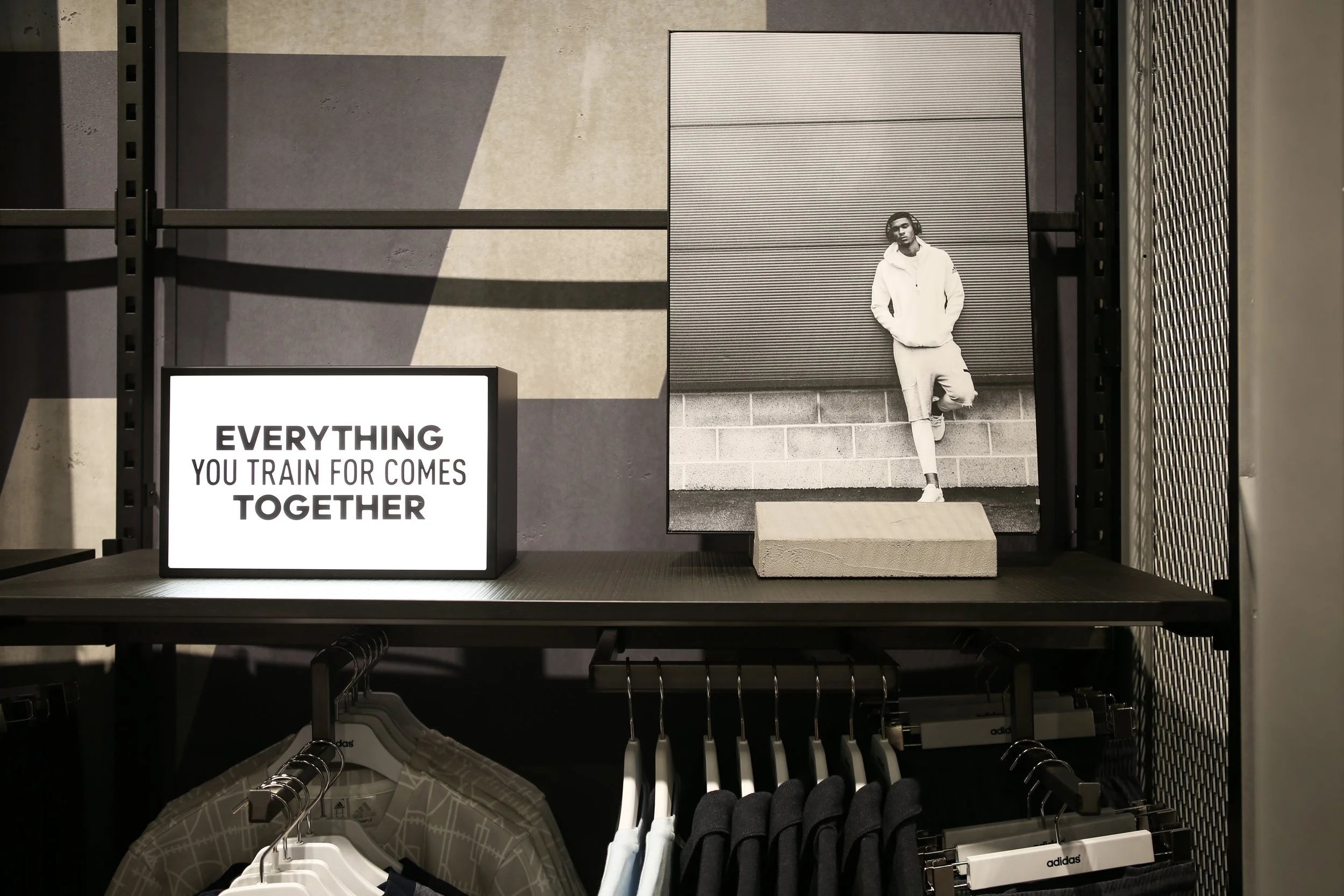 Retail display featuring a large black-and-white photograph of a young man in a hoodie, standing against a wall. A sign in front says, 'EVERYTHING YOU TRAIN FOR COMES TOGETHER'. Below, clothing items are displayed on hangers, including patterned shirts and black garments, with some branded 'adidas'.