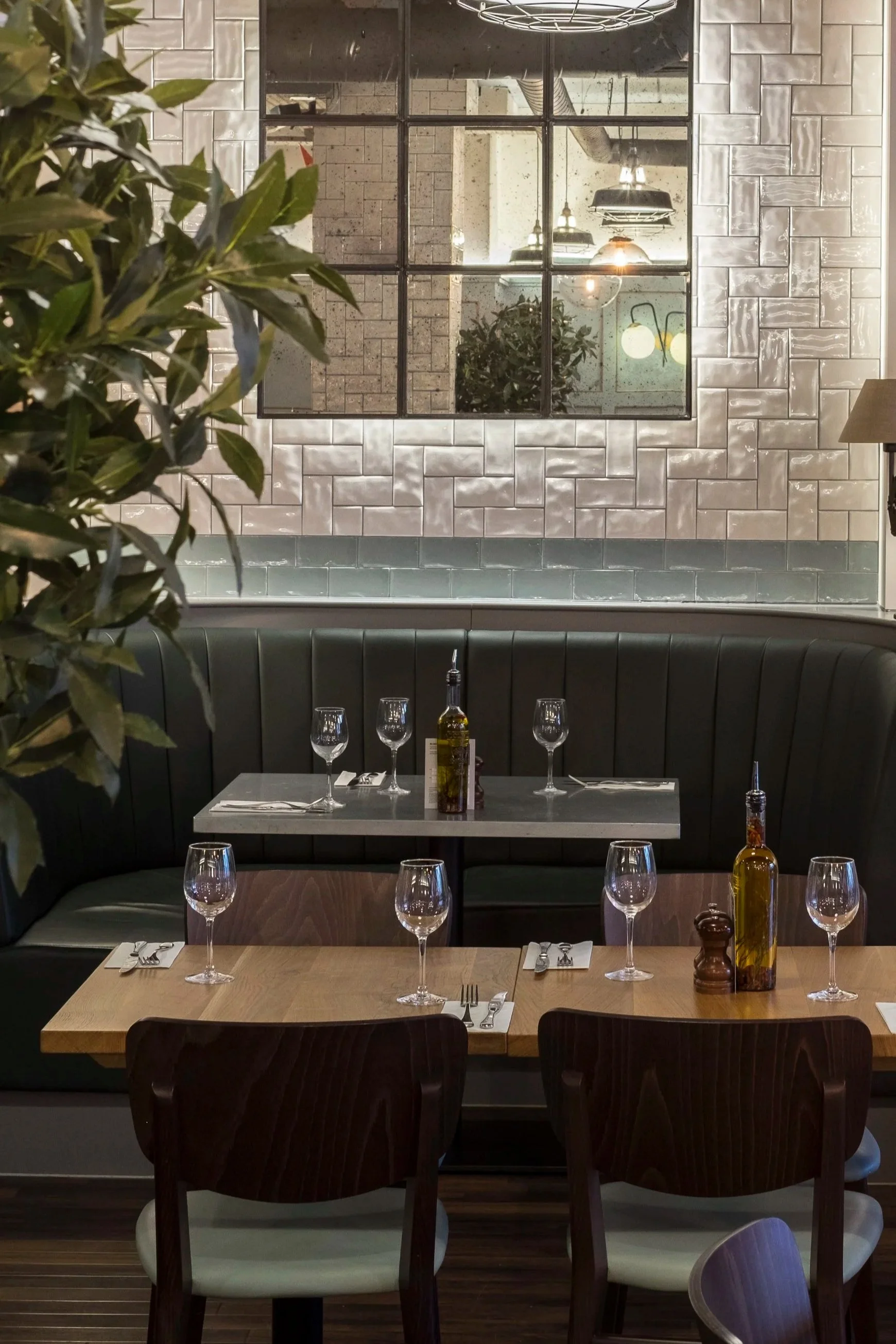 A cozy restaurant scene with a table set for four, including wine glasses, utensils, and bottles of olive oil and pepper. The background features a black leather booth, a white tiled wall, and a large window reflecting industrial-style lighting fixtures and decor.