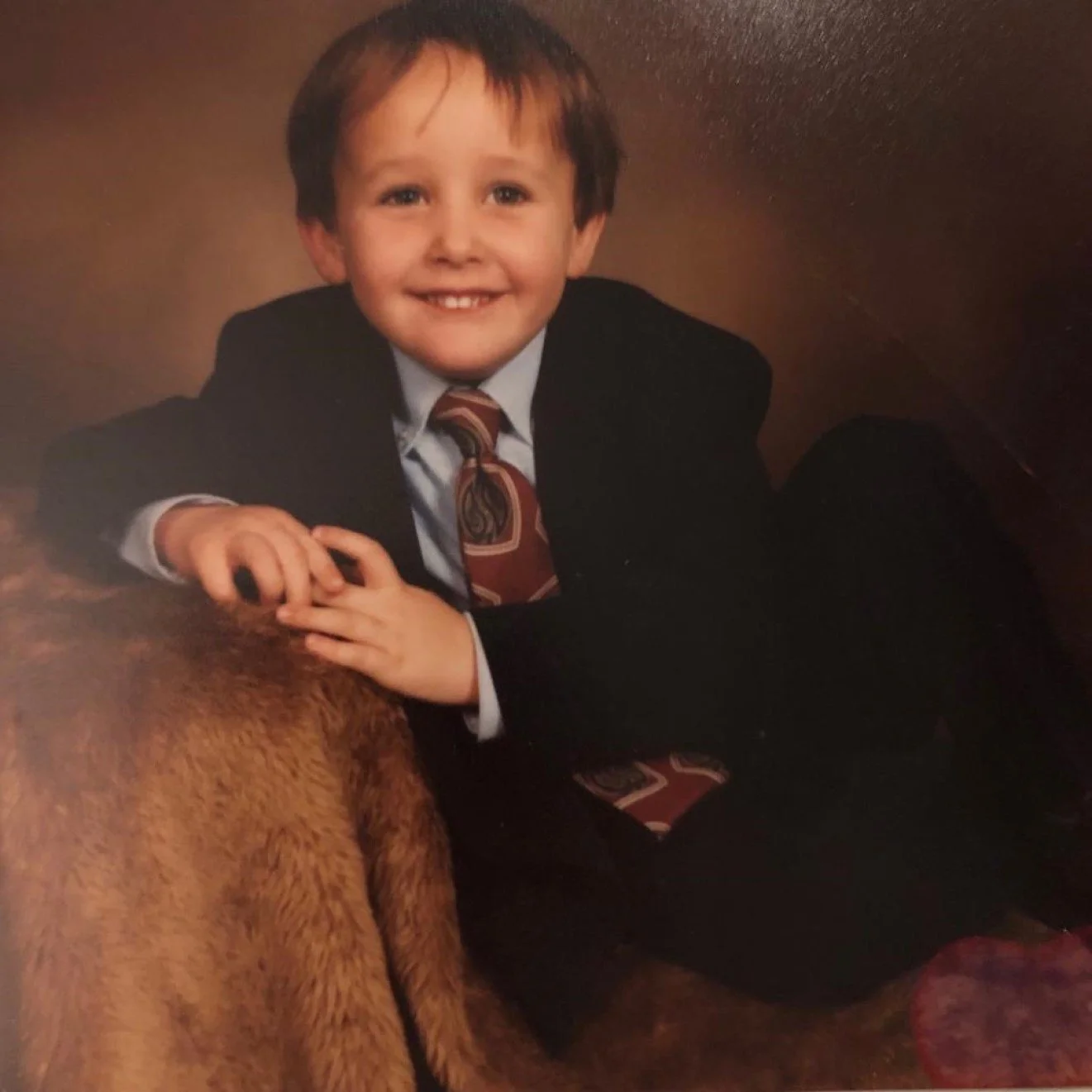 A young boy in a suit and tie sitting on a furry surface, smiling at the camera.