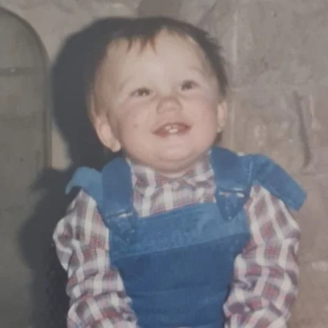 A young child with a big smile, wearing a plaid shirt and denim overalls, in front of a brick wall.