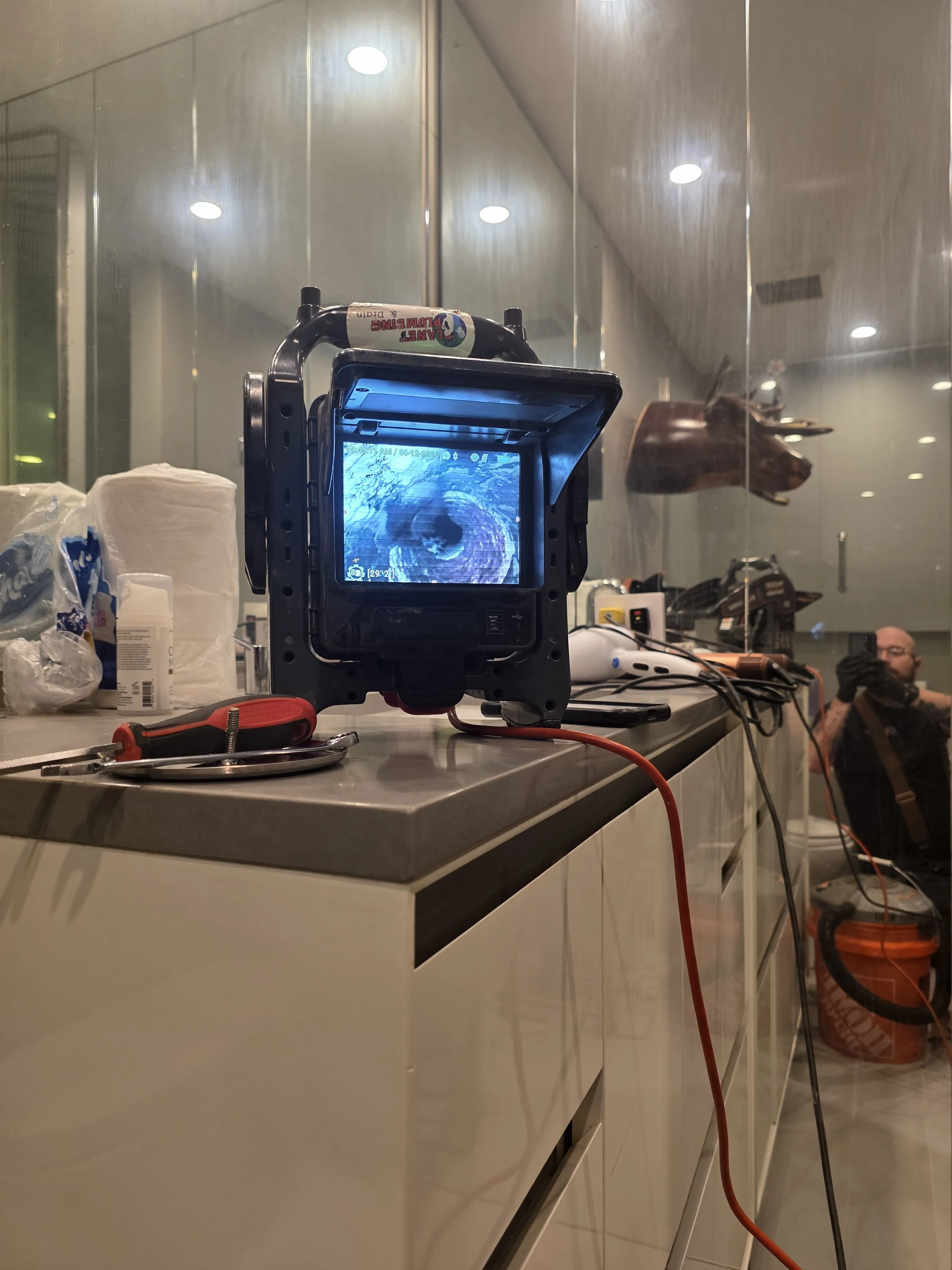 A camera on a countertop in a room with reflective ceiling panels and recessed lighting, with various objects including rolls of paper towels, tools, and a person in the background.