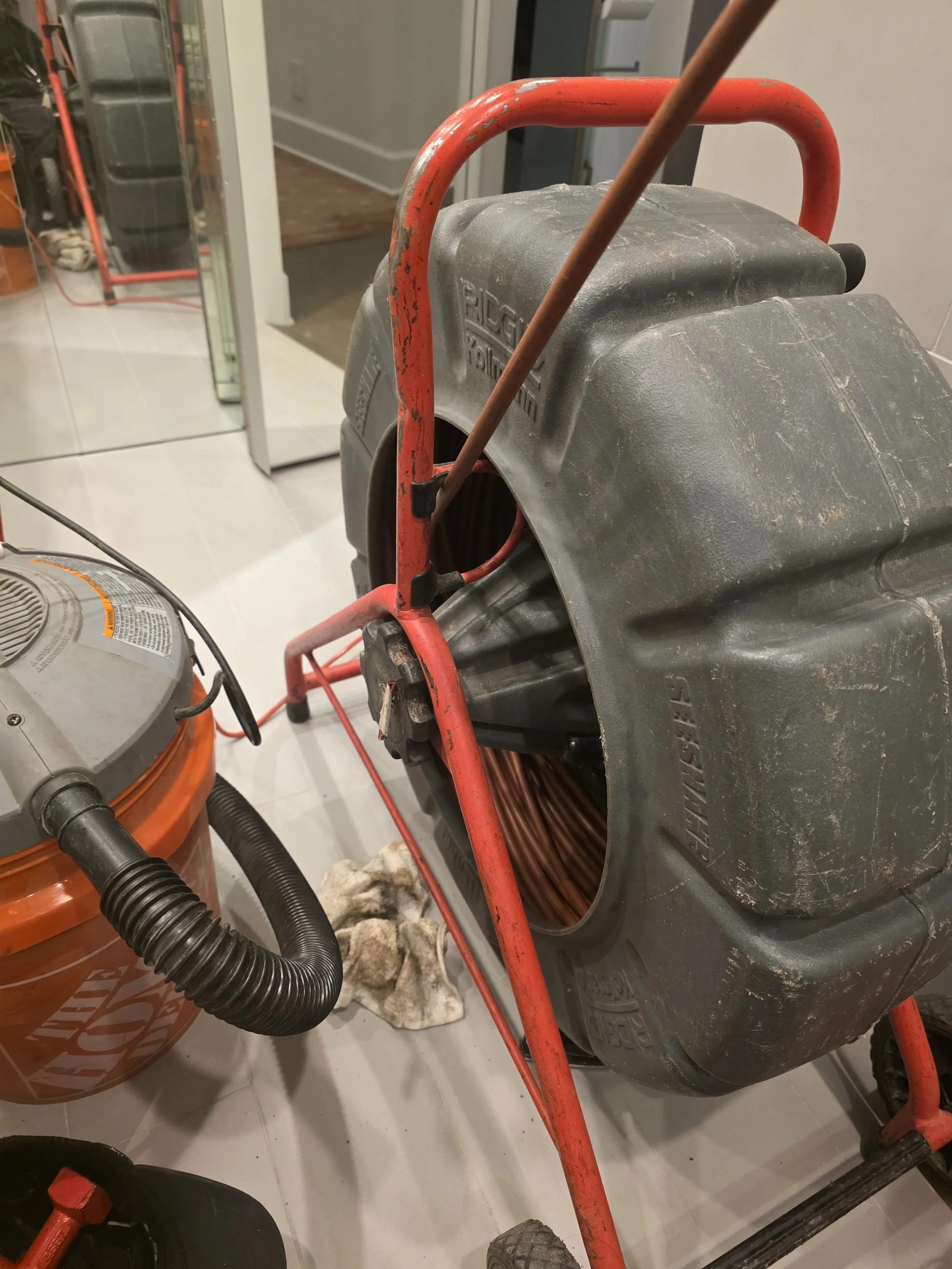 A near view of a concrete cutting saw with a red metal frame, black motor housing, and a large circular blade, positioned on a tiled floor in a workshop or garage.