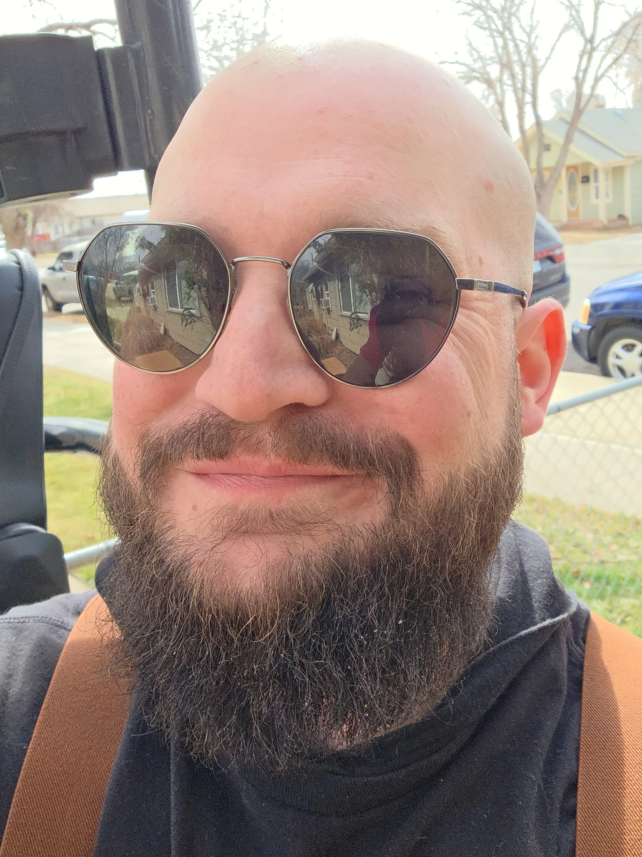 Close-up of a man with a beard wearing sunglasses, a black shirt, and brown straps, standing outdoors with houses and cars in the background.