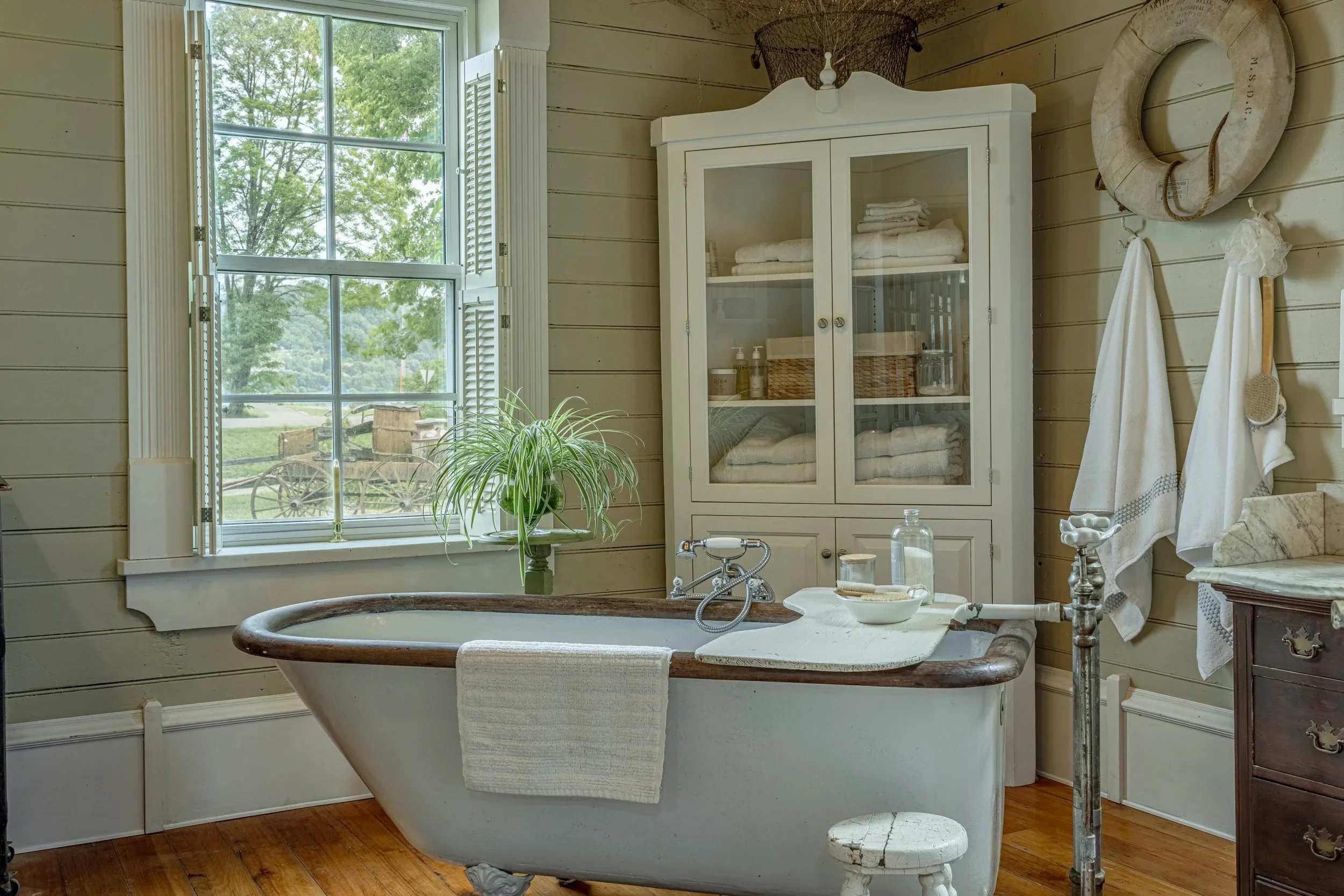 A cozy bathroom with a vintage clawfoot bathtub, filled with water, next to a window with white shutters. There is a green potted plant on the window sill. A white cabinet with towels and toiletries is against the wall. A hanging life preserver is nearby, and there are towels hanging on hooks. The bathroom has wood-paneled walls and hardwood flooring.