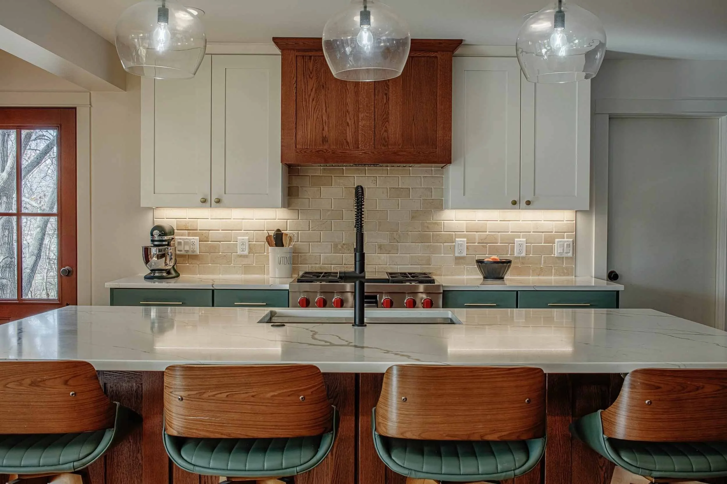 Modern kitchen with white countertops, dark green chairs, white cabinets, a red wooden range hood, beige tile backsplash, and pendant lights.
