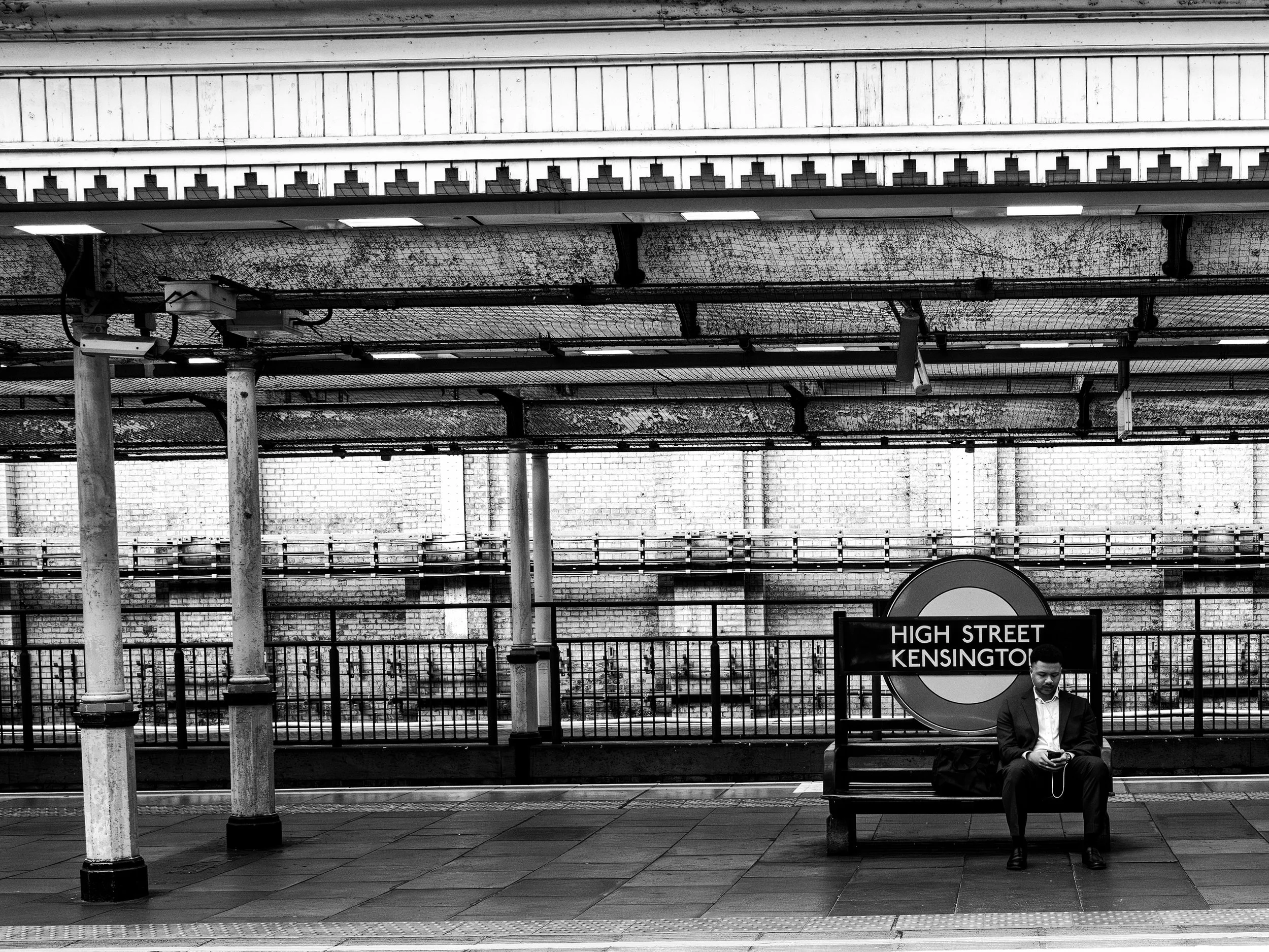 Black and white photo of a bus stop on High Street Kensington with a man sitting on a bench looking at his phone, behind him a fence and brick wall.