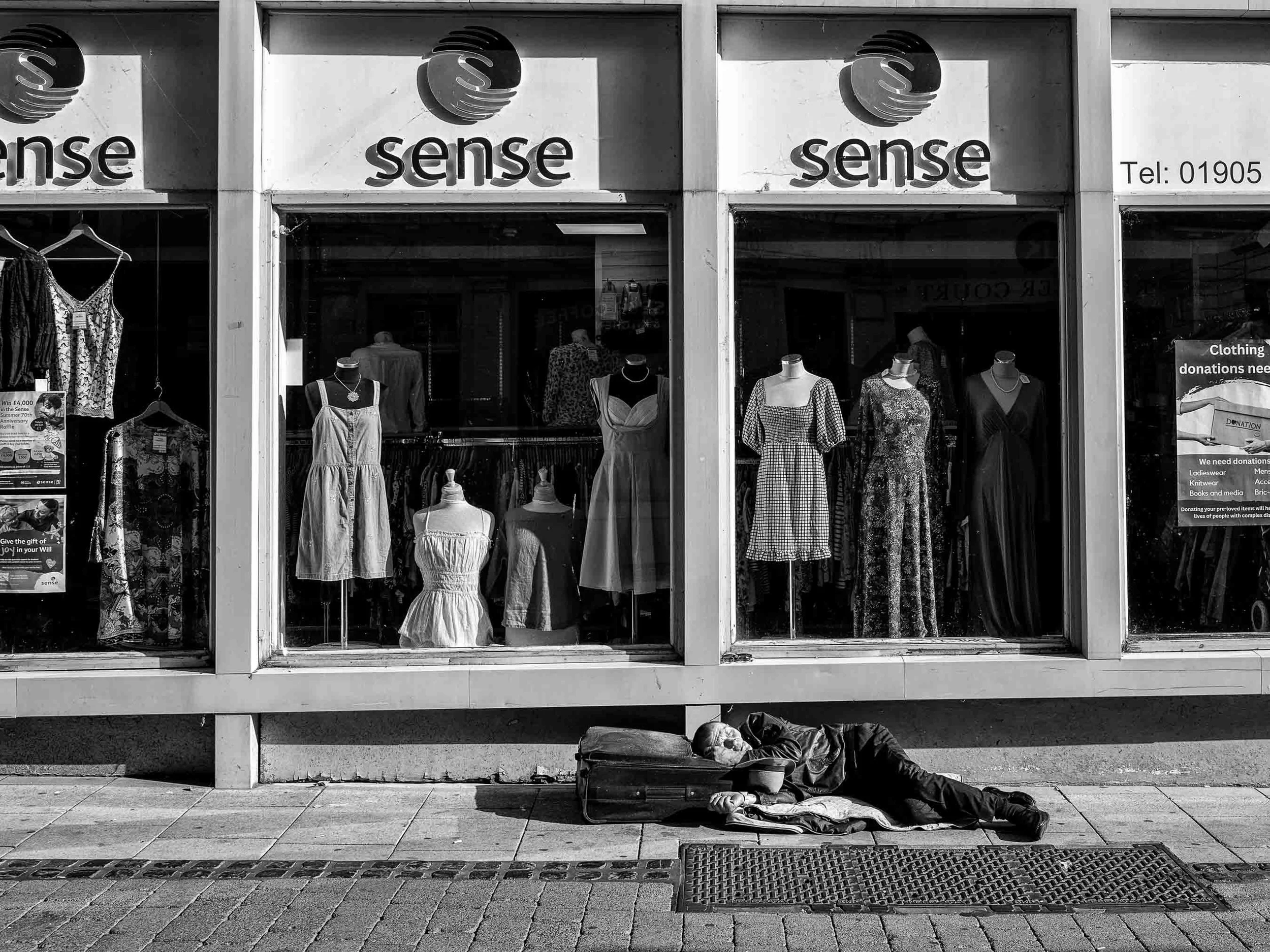 A homeless person sleeping on a sidewalk outside a storefront named 'sense' with mannequins dressed in clothing displayed in the window. The person has a suitcase and wears dark clothing.