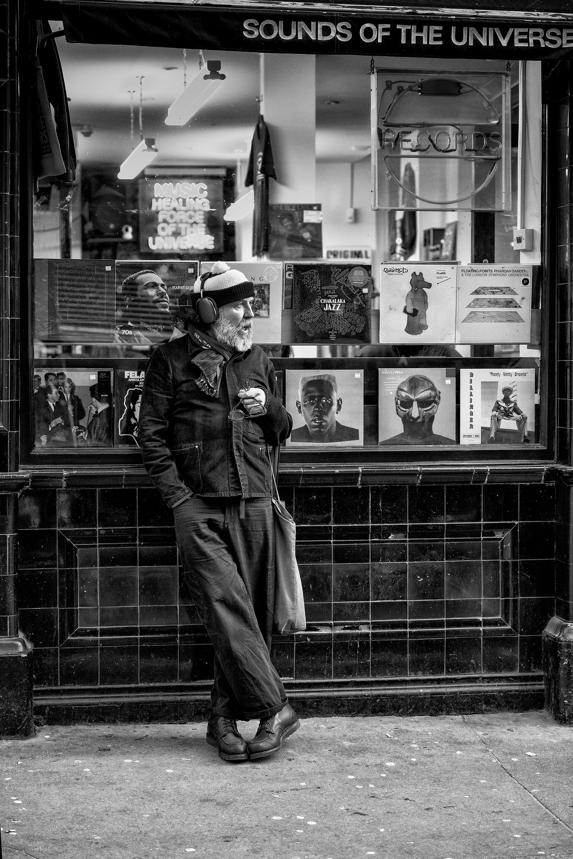 A man with a beard wearing a beanie, headphones, and outdoor clothing standing outside a record shop window, looking to the side, with his hands near his chest, and his legs crossed at the ankles. The shop window displays various vinyl records and posters, with the shop sign reading 'SOUNDS OF THE UNIVERSE' at the top.