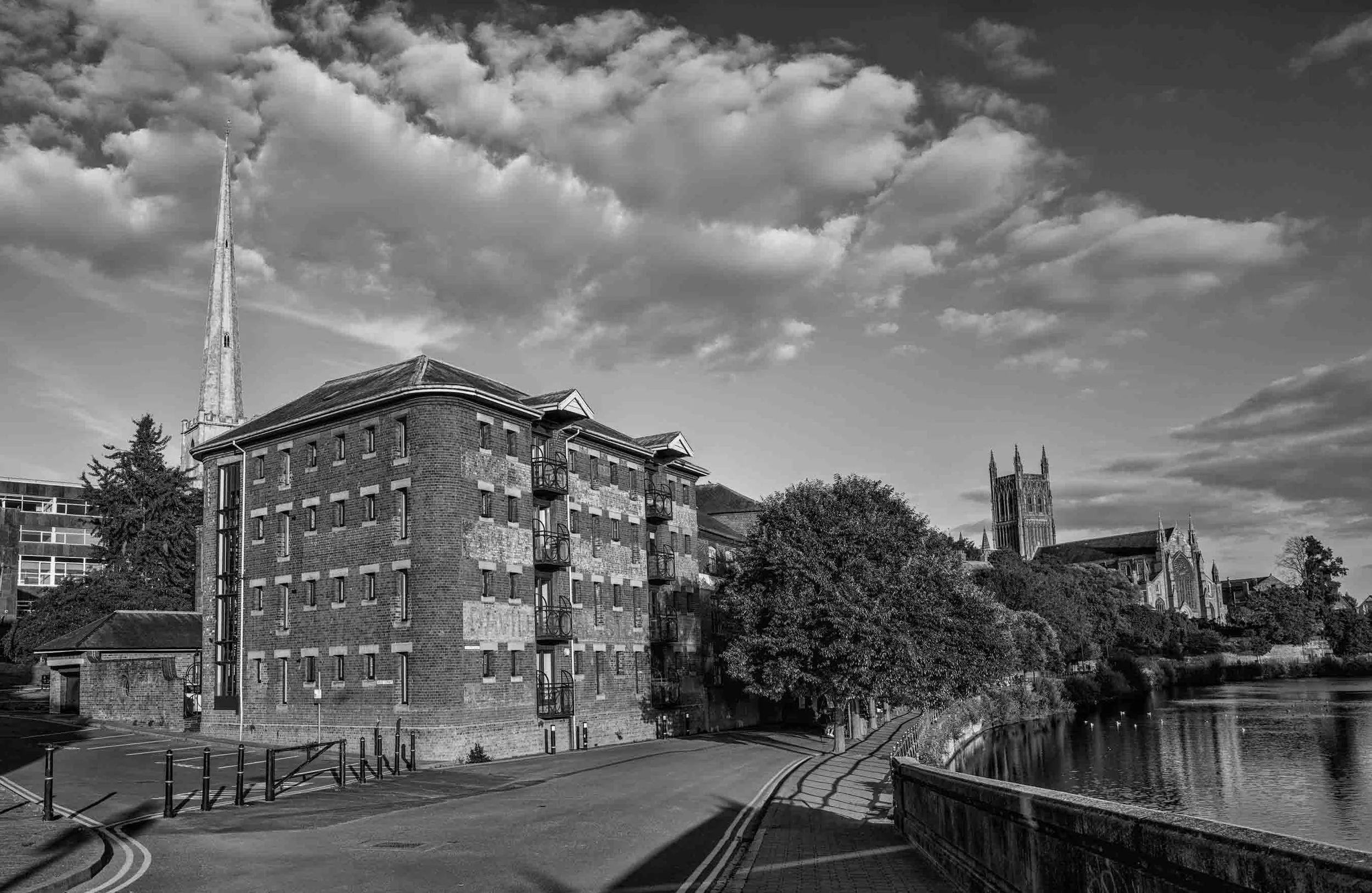 A black and white photo of buildings near a river, with a church tower and a tall spire in the background, cloudy sky, trees, and a curved road in the foreground.