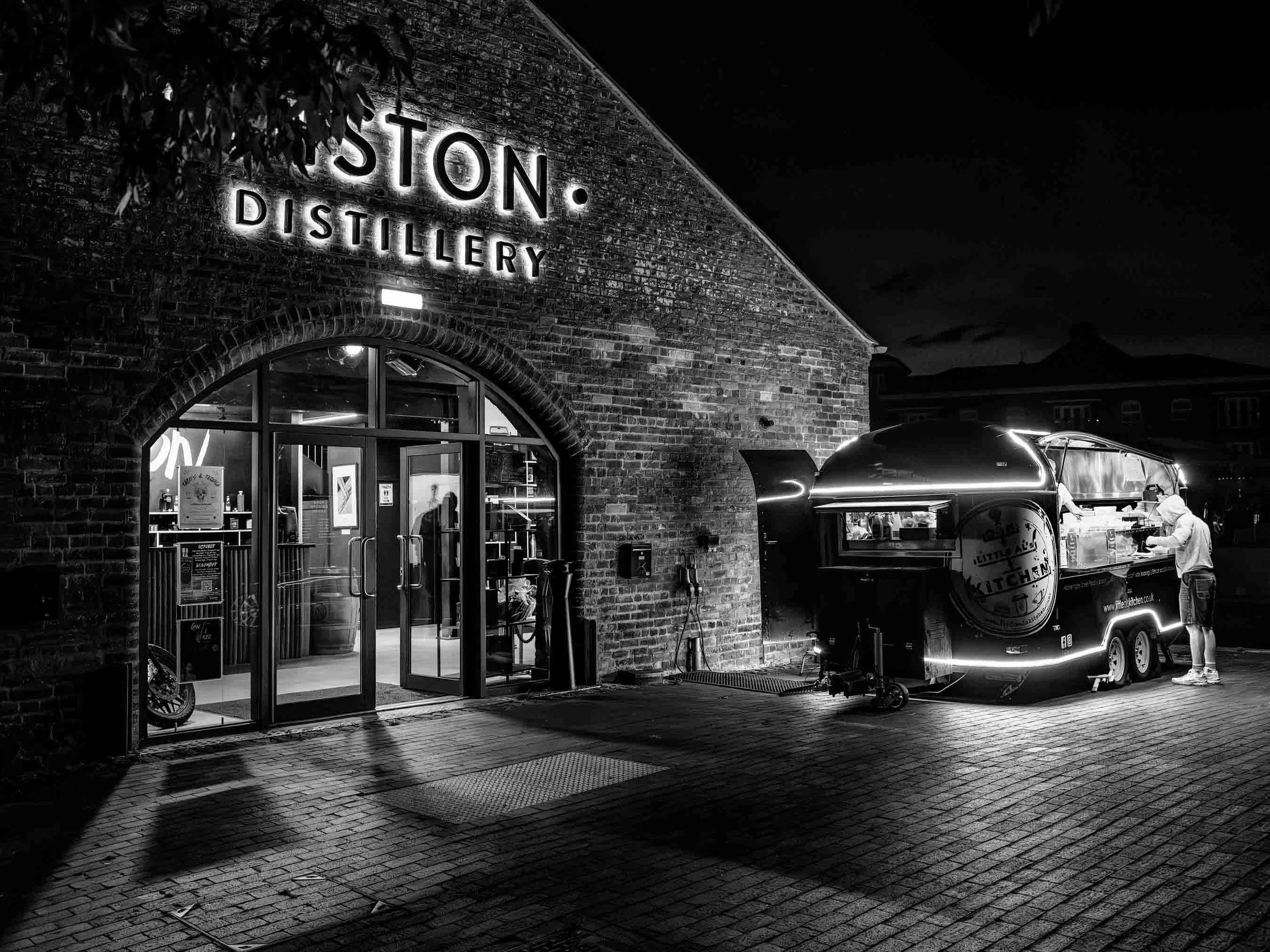 Night scene of a brick building with a sign that says 'STON. DISTILLERY' illuminated. A person is seen near a food truck with a shiny food cart outside. The scene is dark with limited lighting.