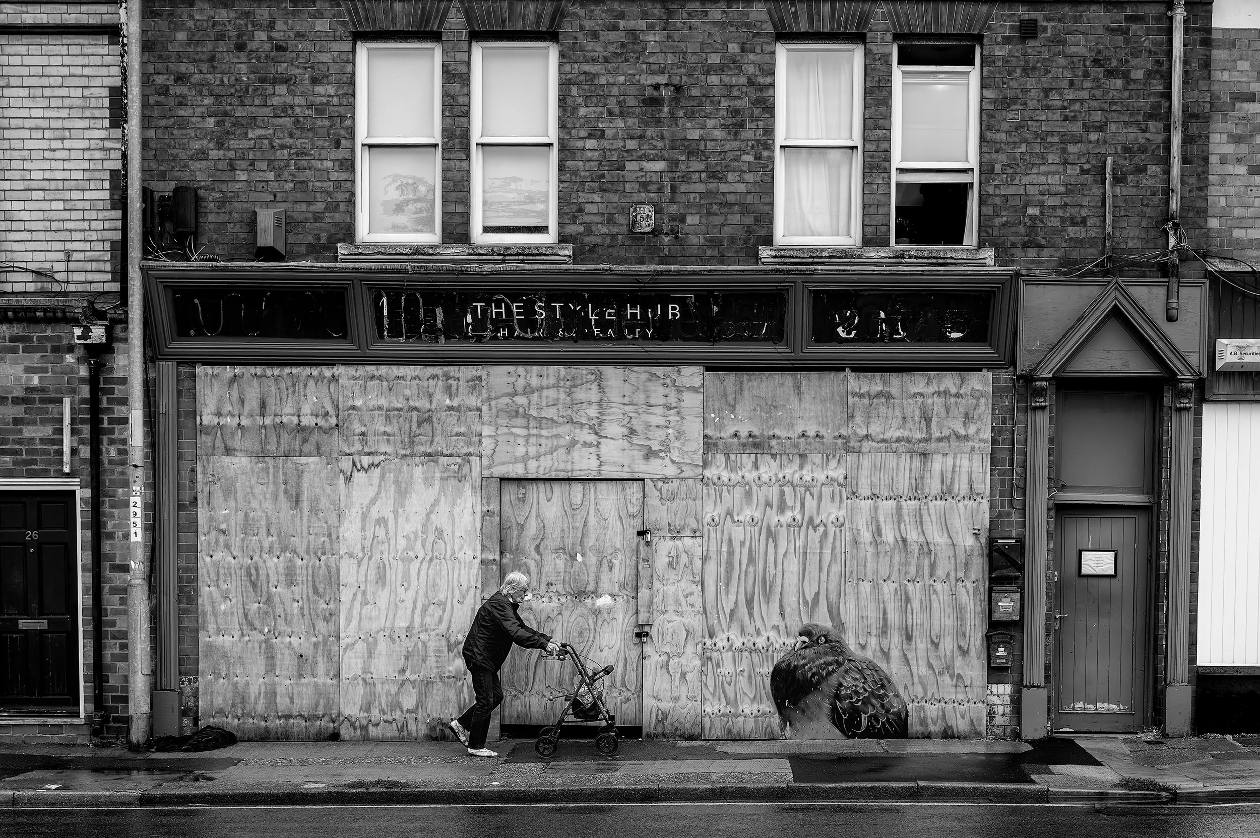 Black and white photo of a person with gray hair riding a bicycle past a boarded-up storefront on a city street, with a person with dark hair in the foreground and a large bird illustration on the right.