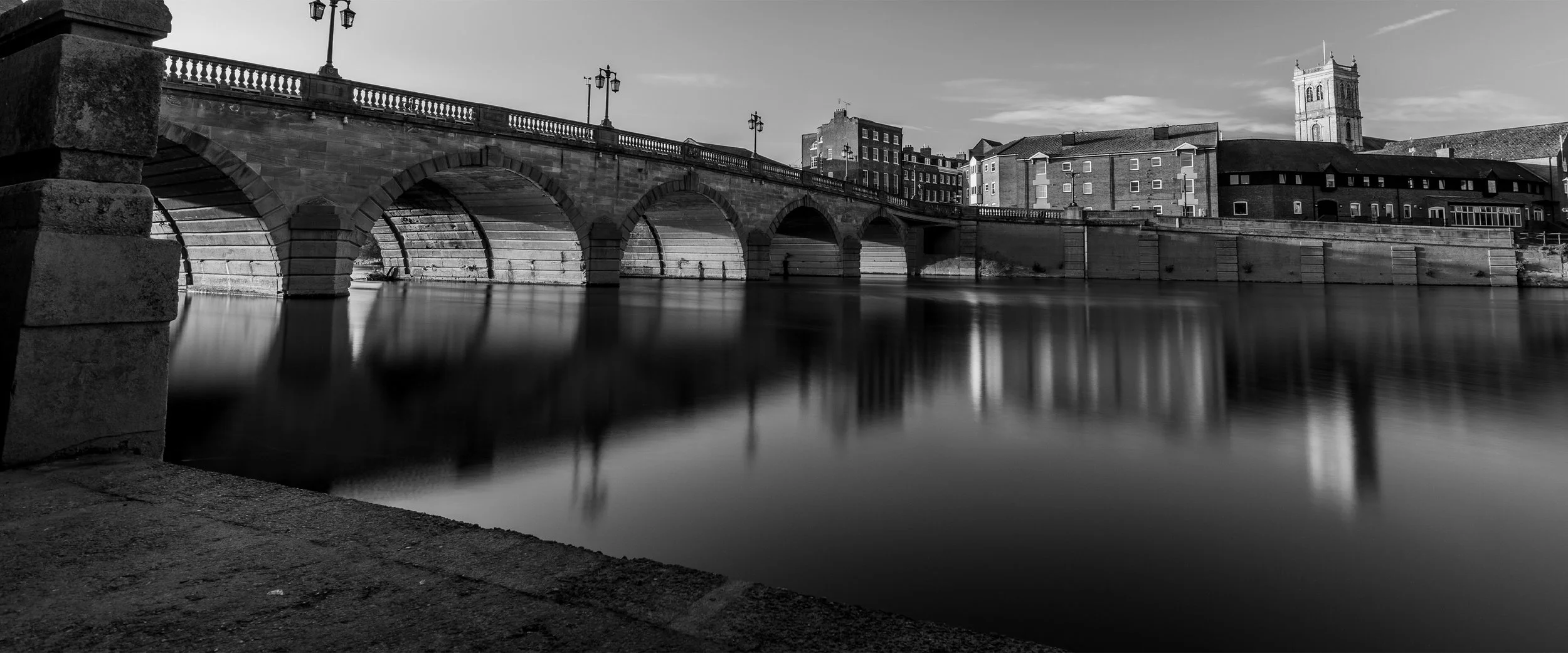 Black and white photo of a stone bridge over a calm river with buildings and a church tower in the background.