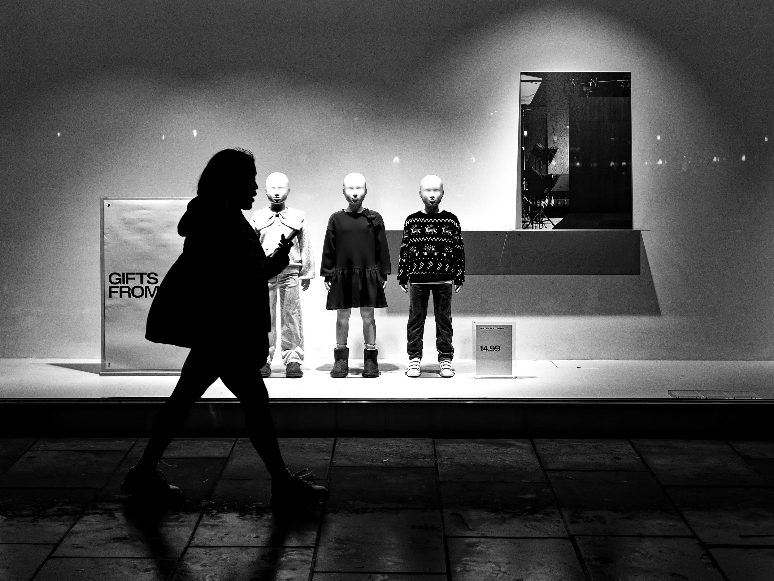 A woman walking past a retail store display window with mannequins dressed in winter clothing, including sweaters and boots, inside a shopping mall.