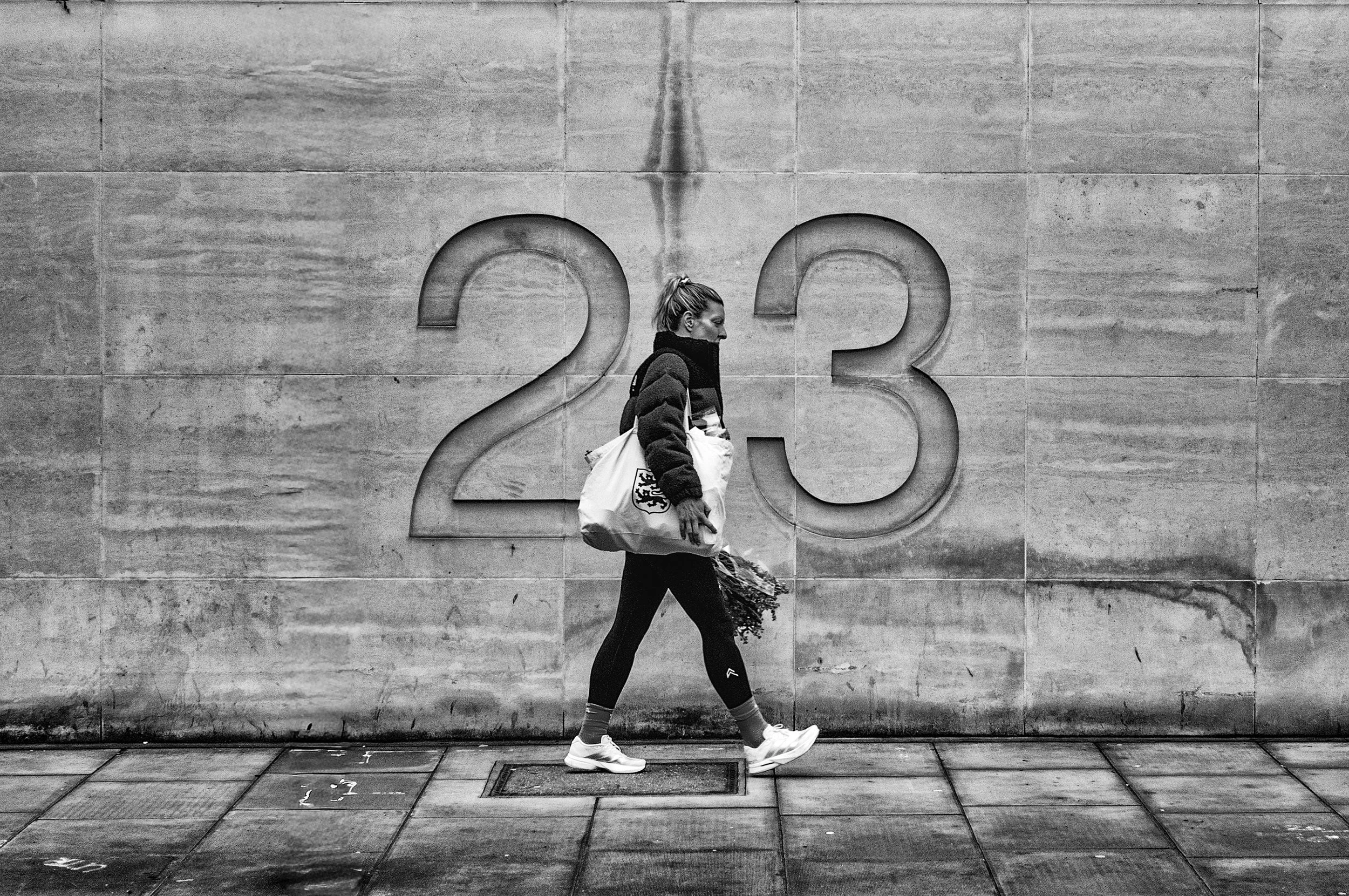 A woman walking past a large wall with the numbers 2 and 3 carved into it, captured in black and white.