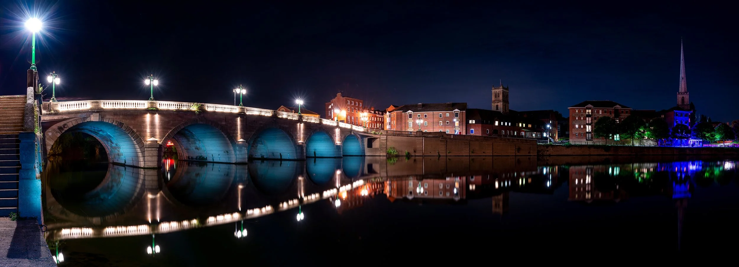 Night view of a cityscape with a bridge over a calm river, illuminated by streetlights, and buildings with lit windows in the background, reflecting colorful lights on the water.