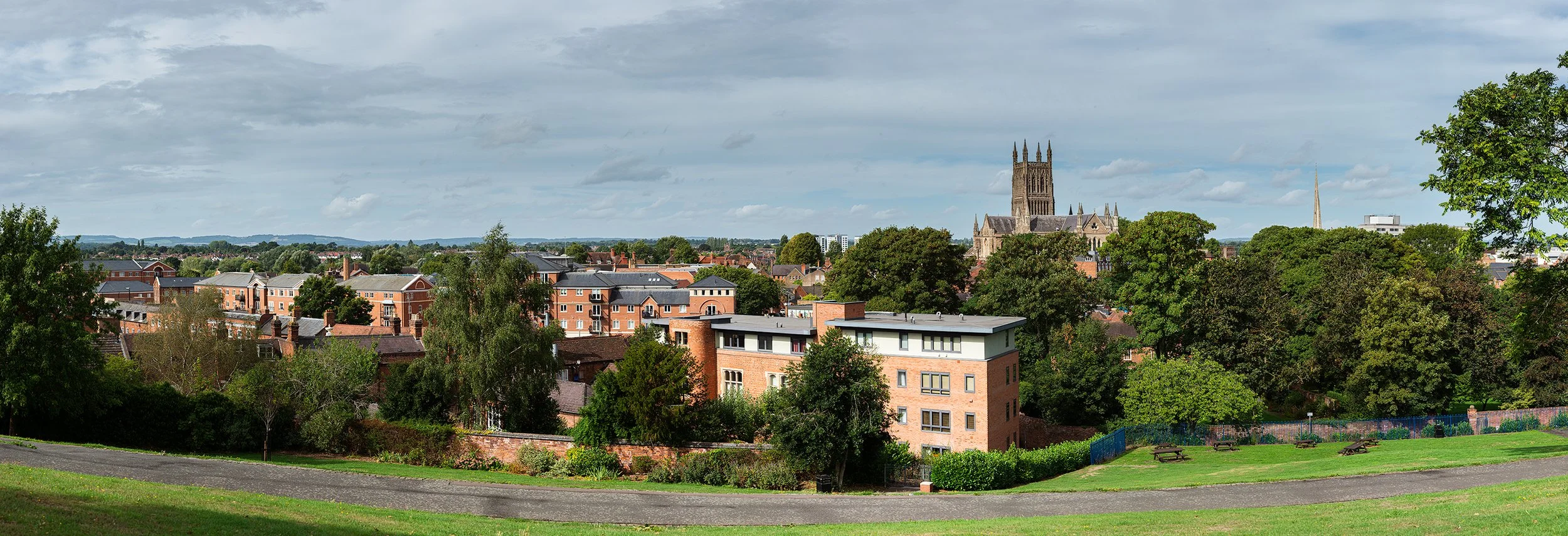 Cityscape with a historic Gothic cathedral, modern apartment buildings, lush green trees, a park with picnic tables, and a partly cloudy sky.