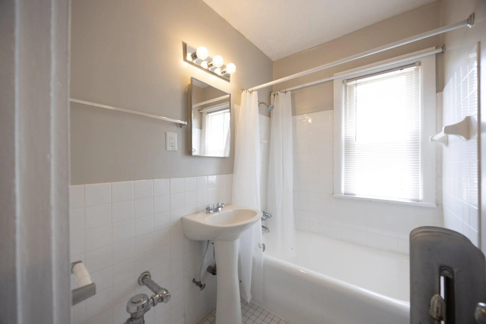 A small bathroom with a sink and a bathtub. The sink has a silver faucet. There is a white curtain on a shower rod above the bathtub. Two windows with blinds are in the background. A rectangular mirror and a light fixture with six bulbs are on the wall.