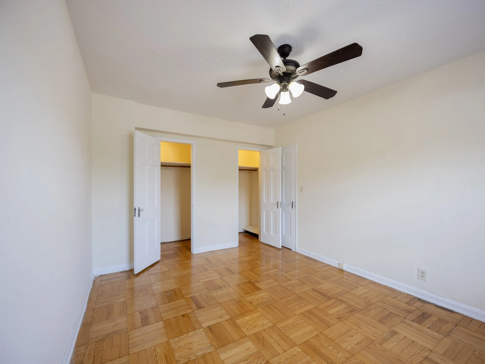 Empty bedroom with hardwood floor, white walls, ceiling fan with lights, and open closet doors.