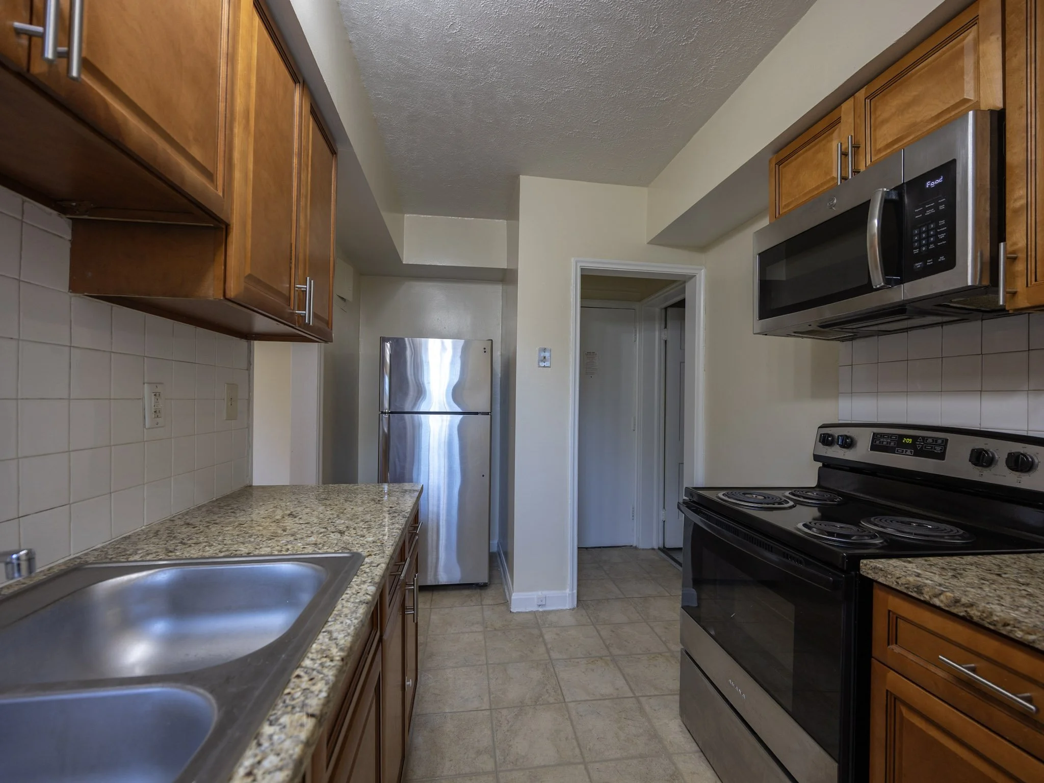 A kitchen with wooden cabinets, granite countertops, a stainless steel refrigerator, a black stove, and a microwave, with beige tiled floors and a white tiled backsplash.