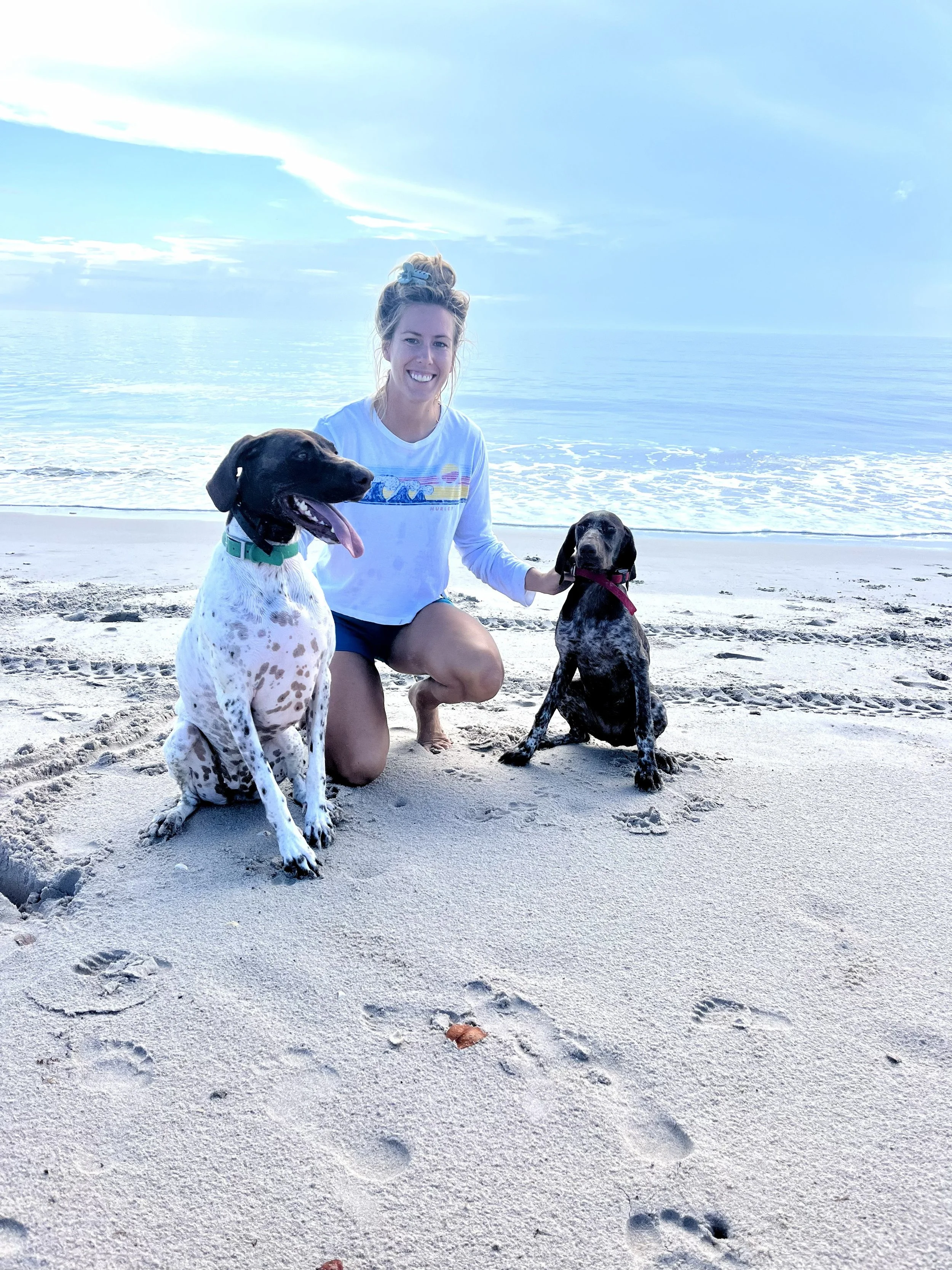 A woman with blonde hair tied up in a bun, wearing a white long-sleeve shirt with a colorful wave design, kneeling on a sandy beach with two dogs, one black and white and the other black and brindle, near the water with ocean waves and a blue sky in the background.