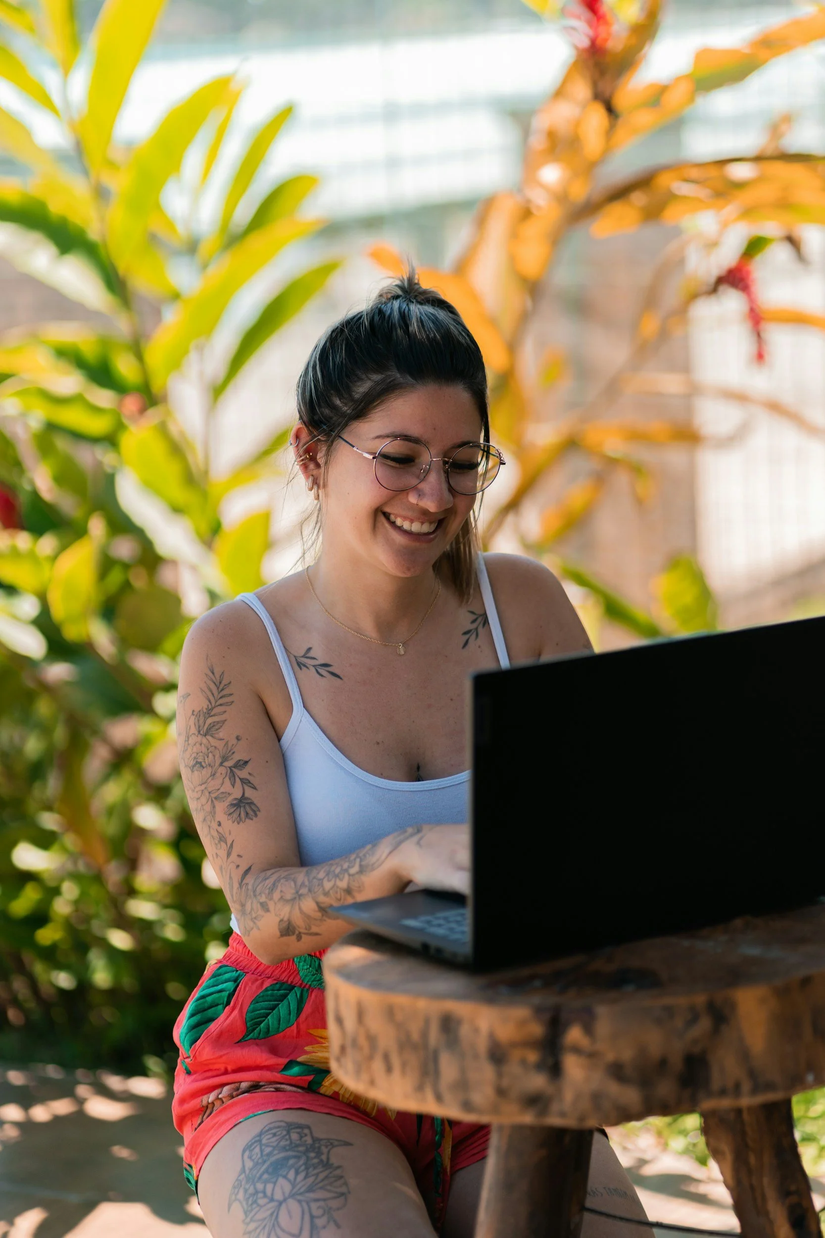 A woman with black hair tied up, wearing round glasses and a white tank top, smiling while working on a laptop outdoors. She has tattoos on her arm and thigh, and is seated at a rustic round wooden table in a lush, green environment with large leafy plants in the background.