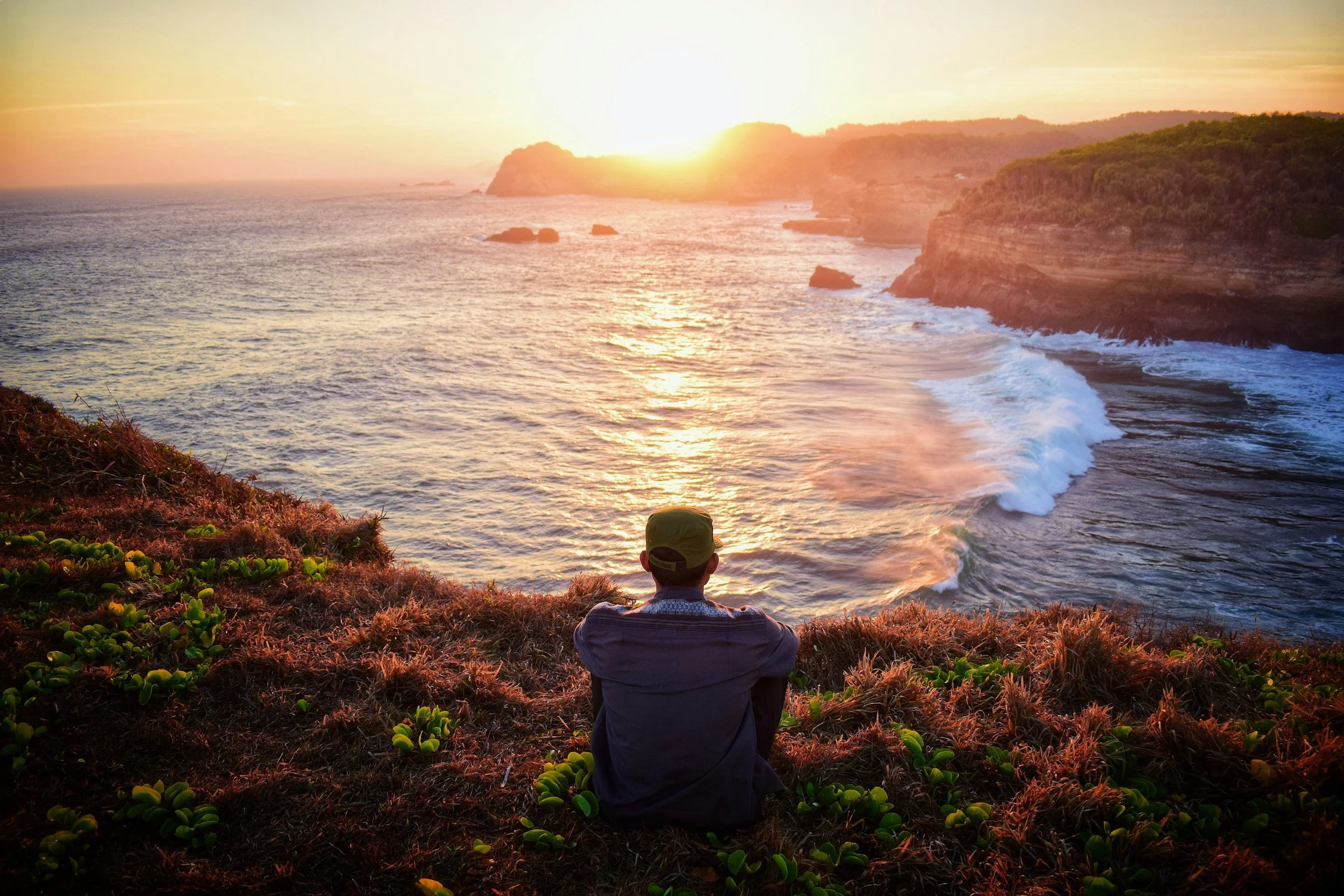 Person sitting on a grassy hill overlooking the ocean at sunset, with cliffs in the background.