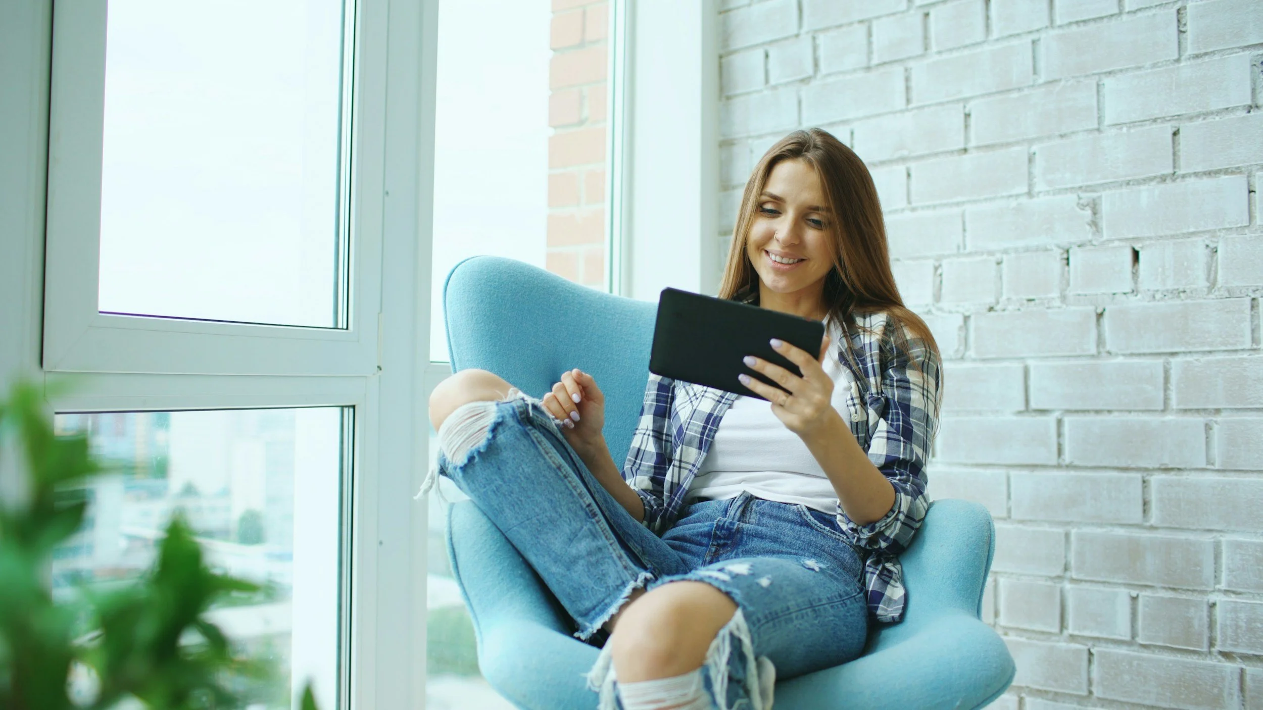 A young woman with long hair, wearing a plaid shirt and ripped jeans, sitting on a blue armchair near a large window with a brick wall behind her, smiling while looking at her phone.