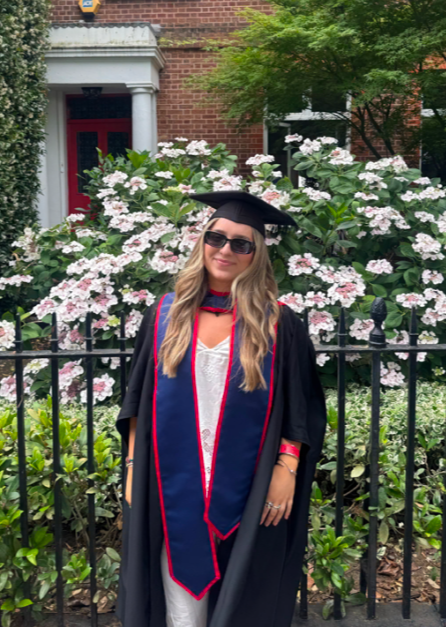 A young woman in graduation gown and cap standing outdoors in front of blooming pink and white flowers with a brick building and green foliage in the background.