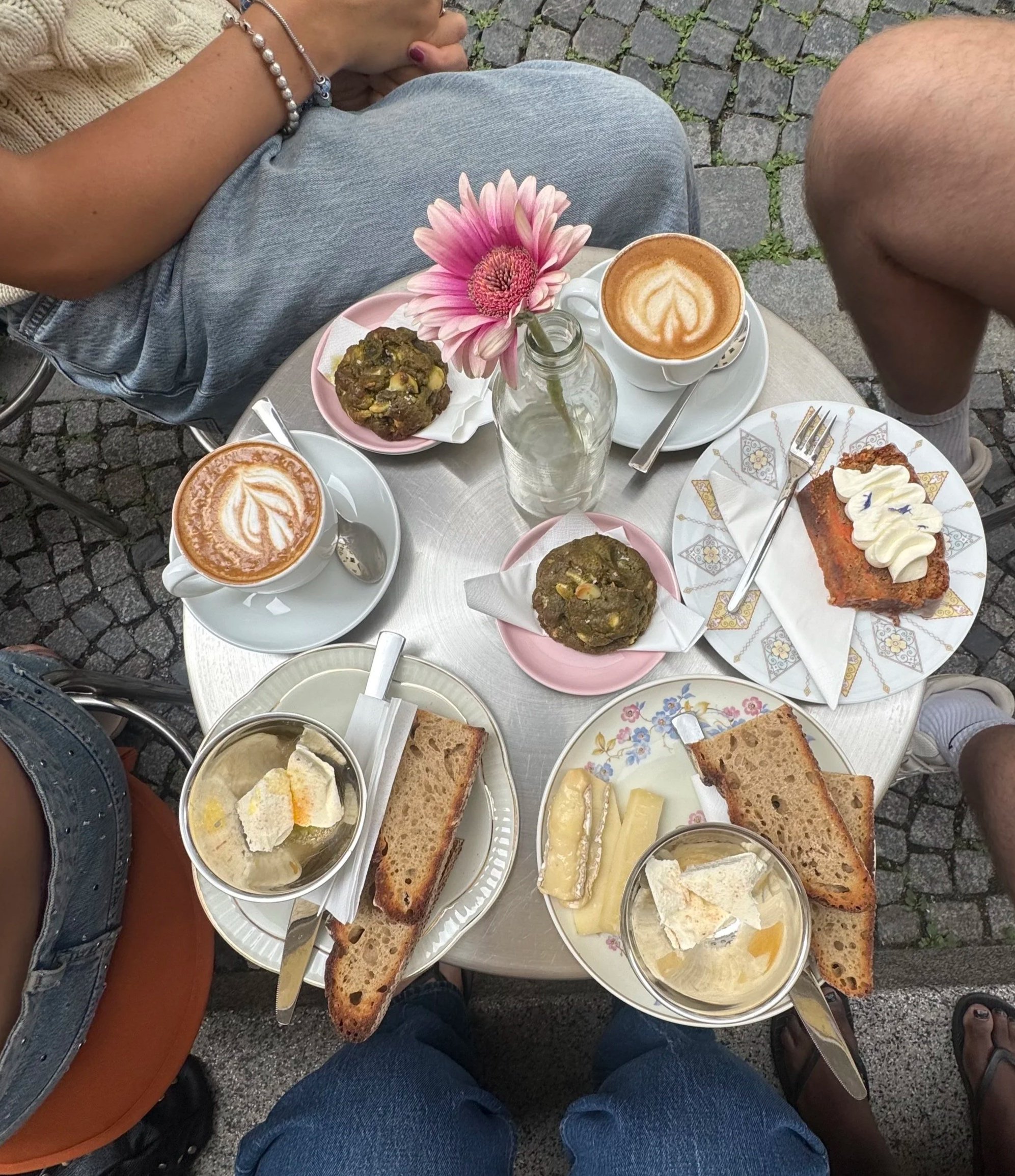 Overhead view of a cafe table with cups of coffee with latte art, slices of bread, a small jar with pink flowers, and cookies on pink plates, with people sitting around on a cobblestone outdoor setting.