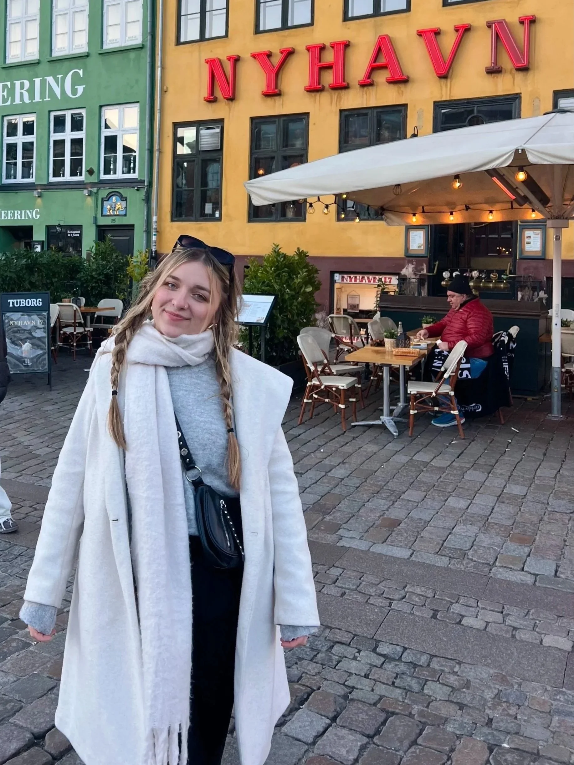 A young woman with long blonde hair in braids, wearing a grey sweater, a white scarf, and a long light-colored coat, smiling at the camera. Behind her, there is an outdoor café with white chairs, tables, a large white umbrella, and a man in a red jacket and black beanie sitting at one of the tables. The background features colorful buildings, one with yellow walls and large windows, and another with green walls, with neon signs reading 'NYHAVN' and 'BREWING'.