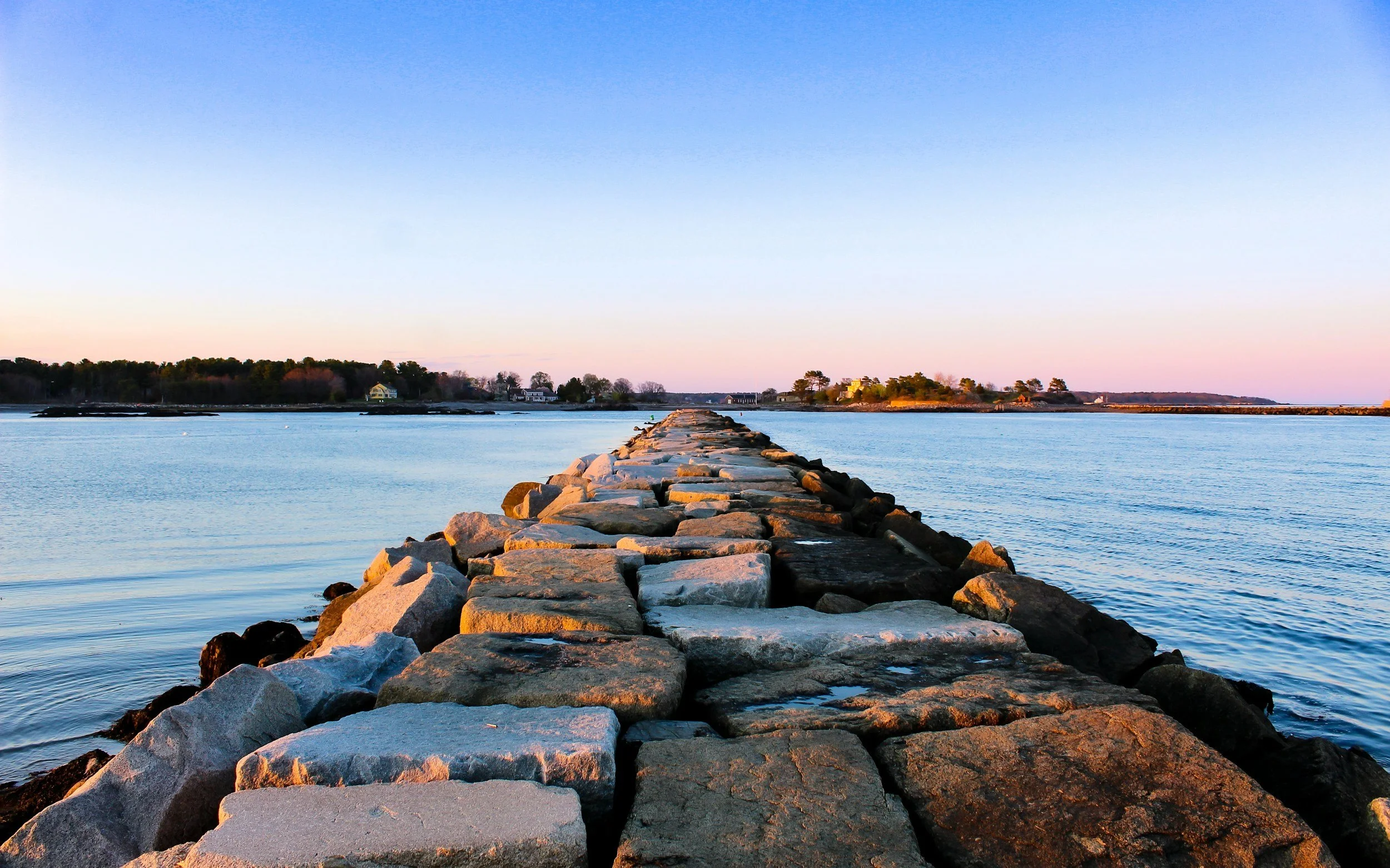 A stone jetty extending into calm water with trees and houses visible on the shoreline in the distance, under a clear sky at sunset.