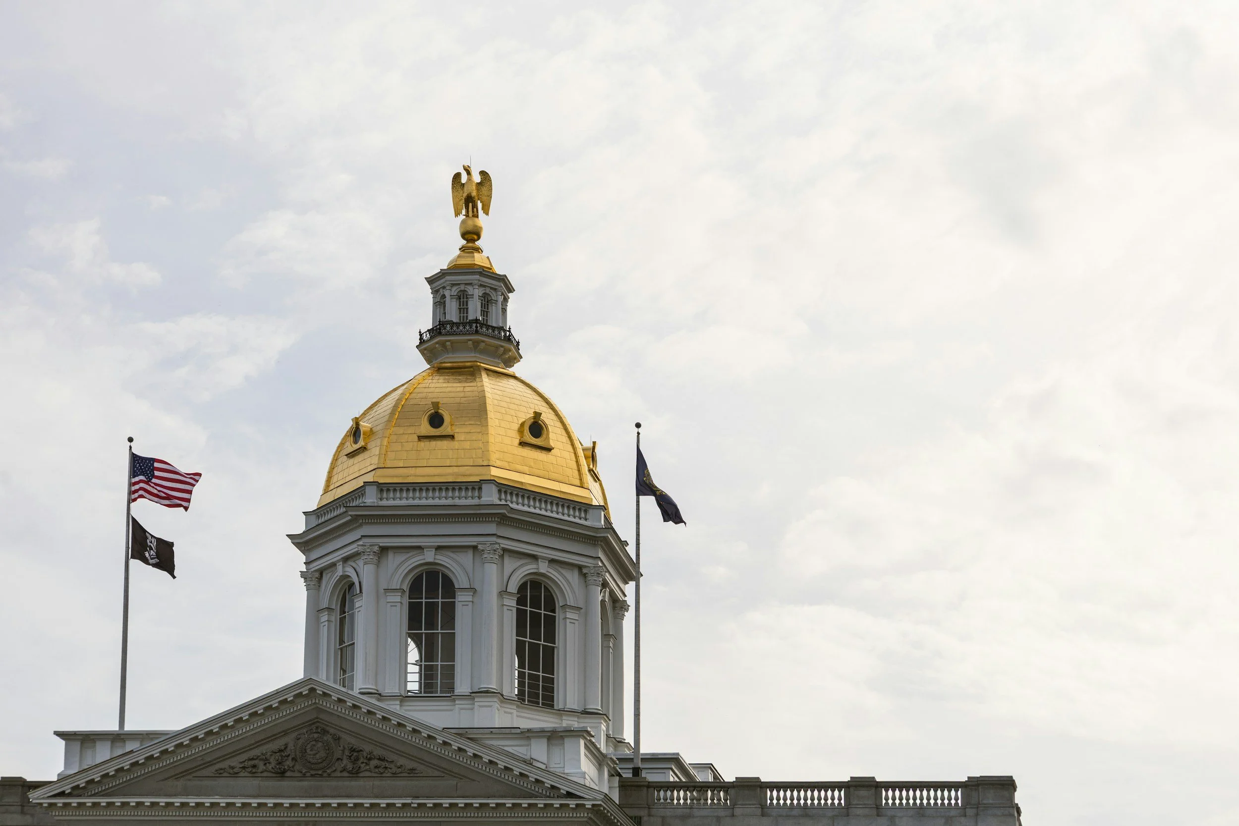 A building with a gold dome and a statue of an eagle on top, featuring flags of the United States and other entities nearby, under a partly cloudy sky.