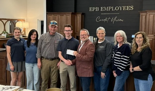 Eight people standing together inside an EPB EMPLOYEES Credit Union, with two men in the center shaking hands and exchanging a check.