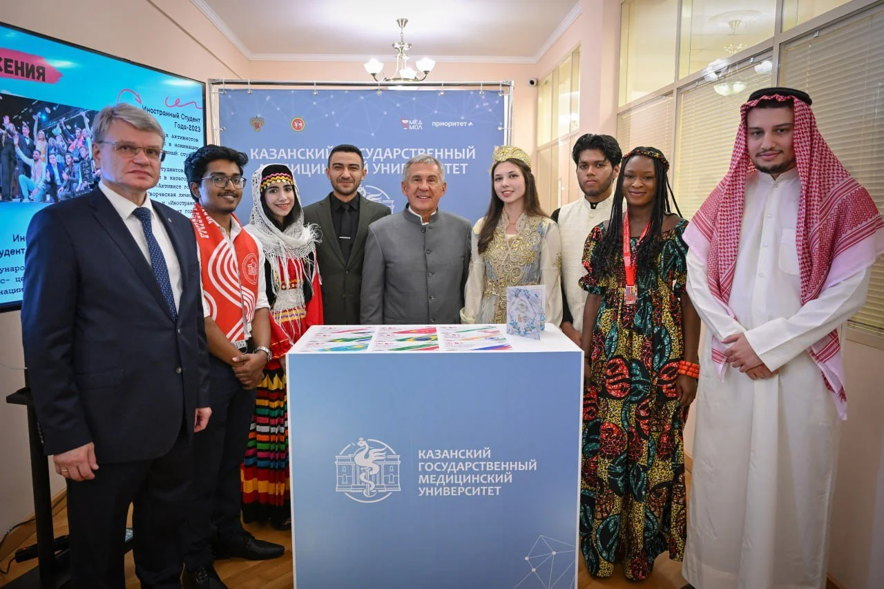 Group of diverse students and officials standing behind a university booth, with some dressed in traditional clothing and others in formal attire, at the Kazan State Medical University event.